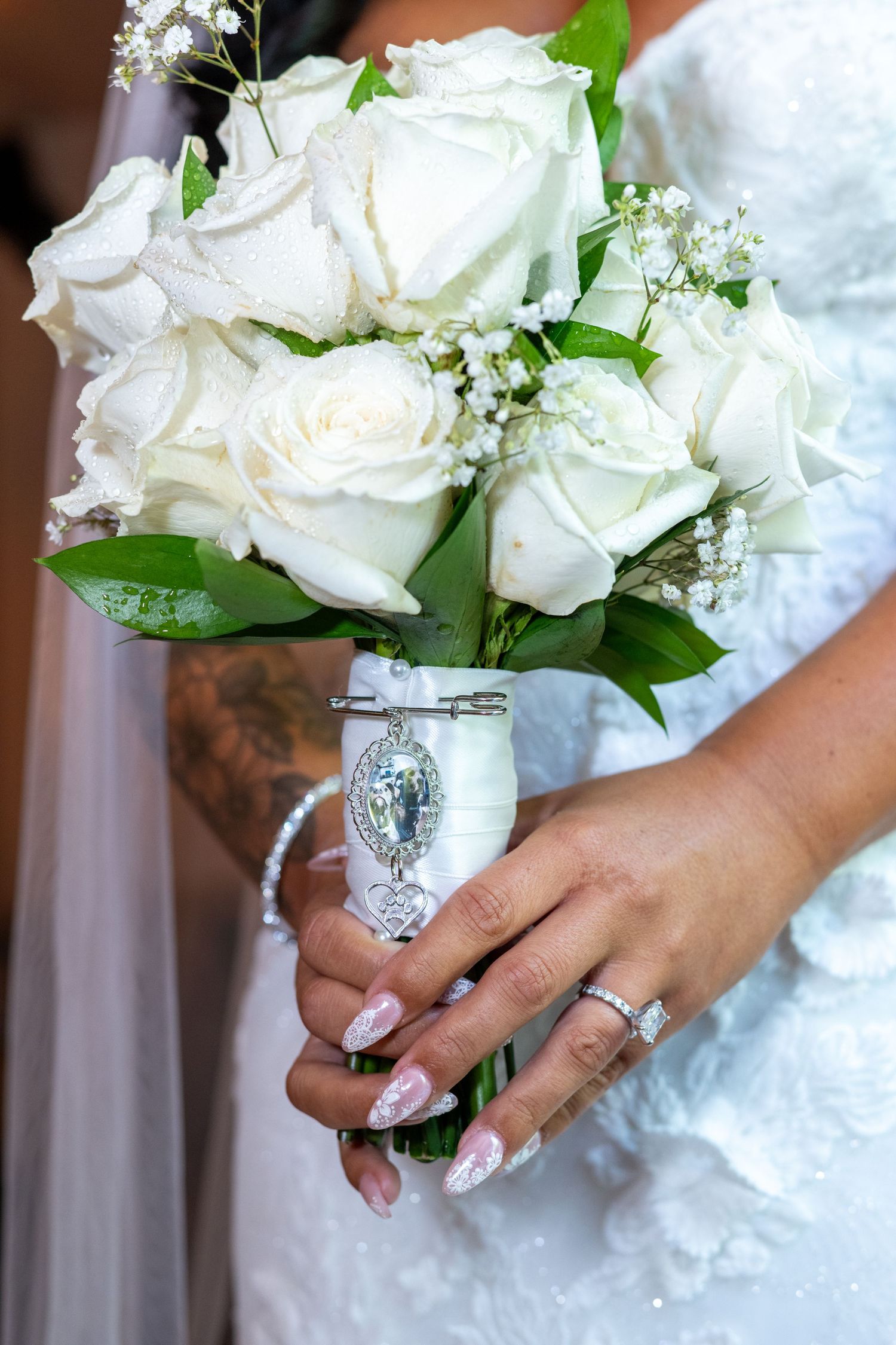 Bride holding a white rose wedding bouquet with silver brooch, wearing a diamond ring and white lace gown.