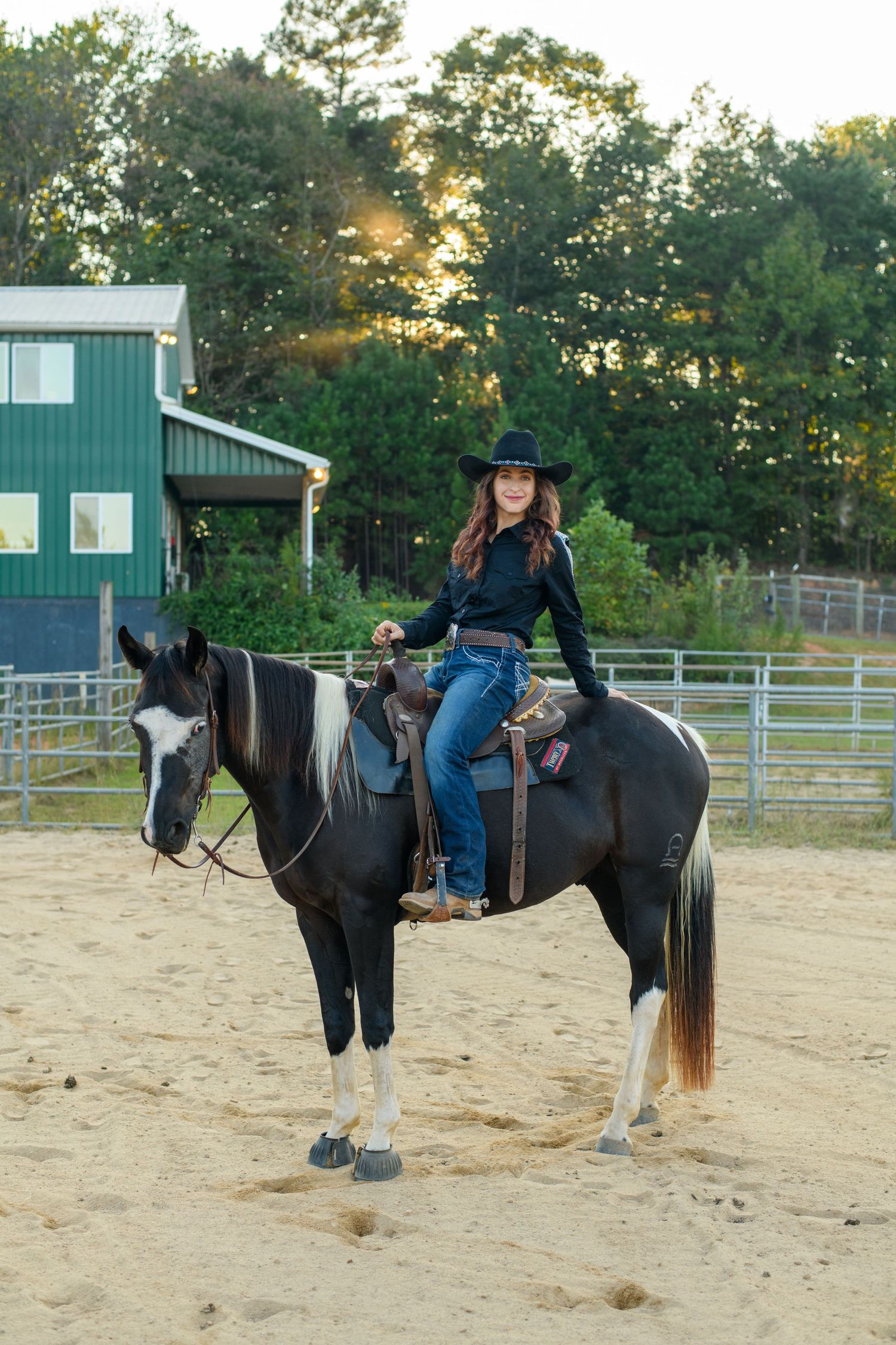 A smiling woman in a black cowboy hat and jeans rides a black and white paint horse in a sandy arena.