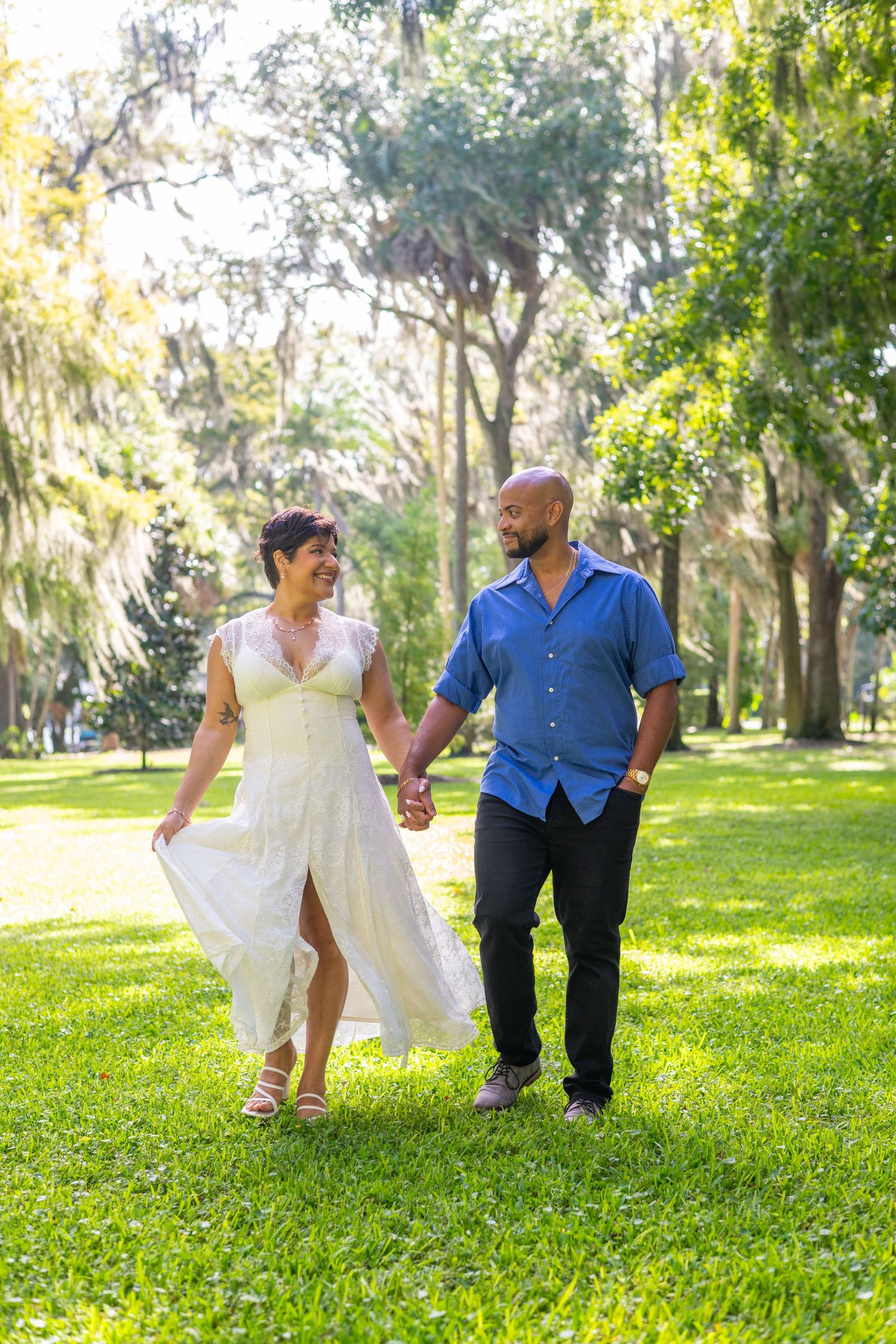 A couple holds hands walking through a sunlit park, woman in white dress, man in blue shirt, lush green grass.