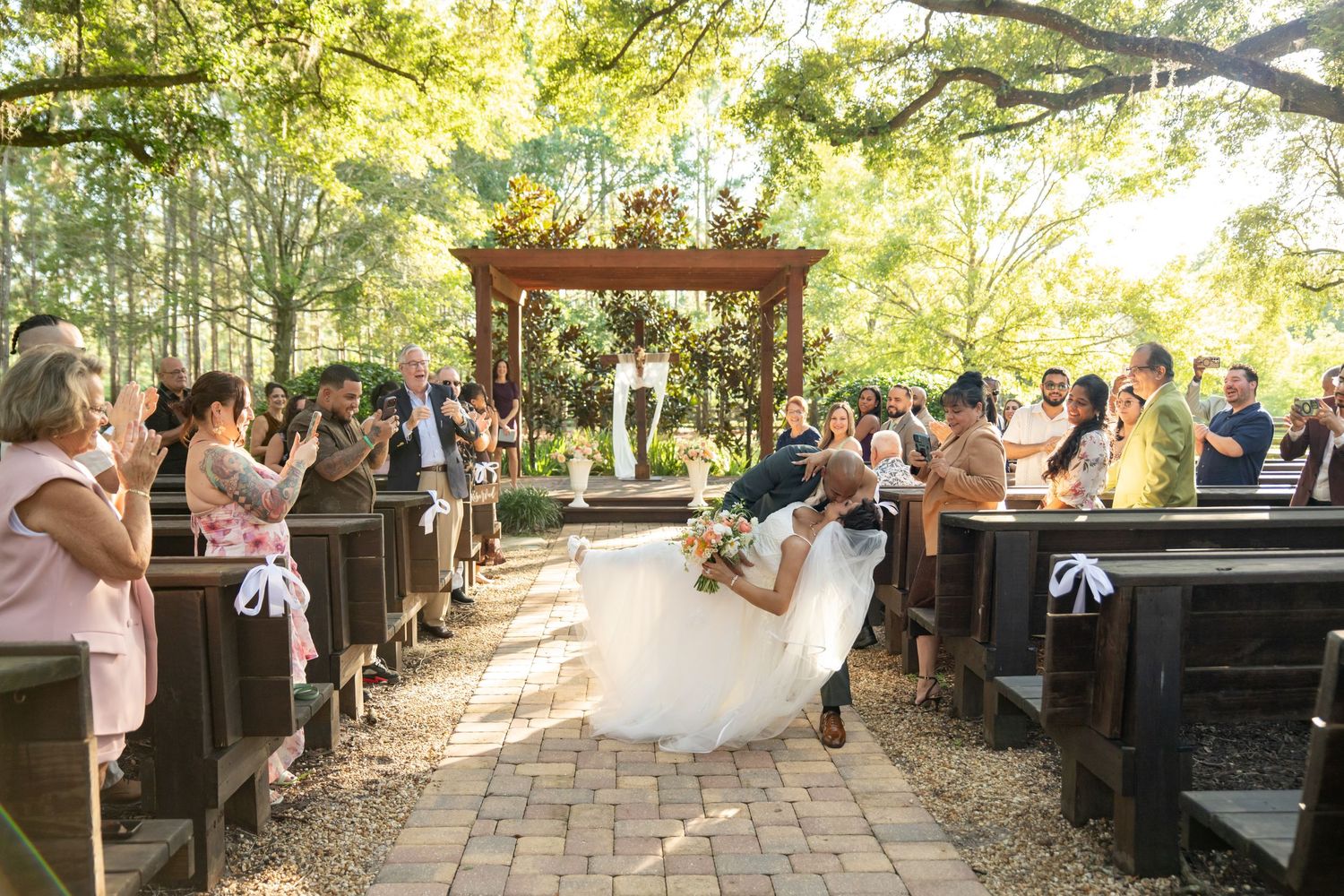 Bride and groom share a romantic dip kiss at outdoor wedding ceremony with wooden arbor and guests seated on pews.