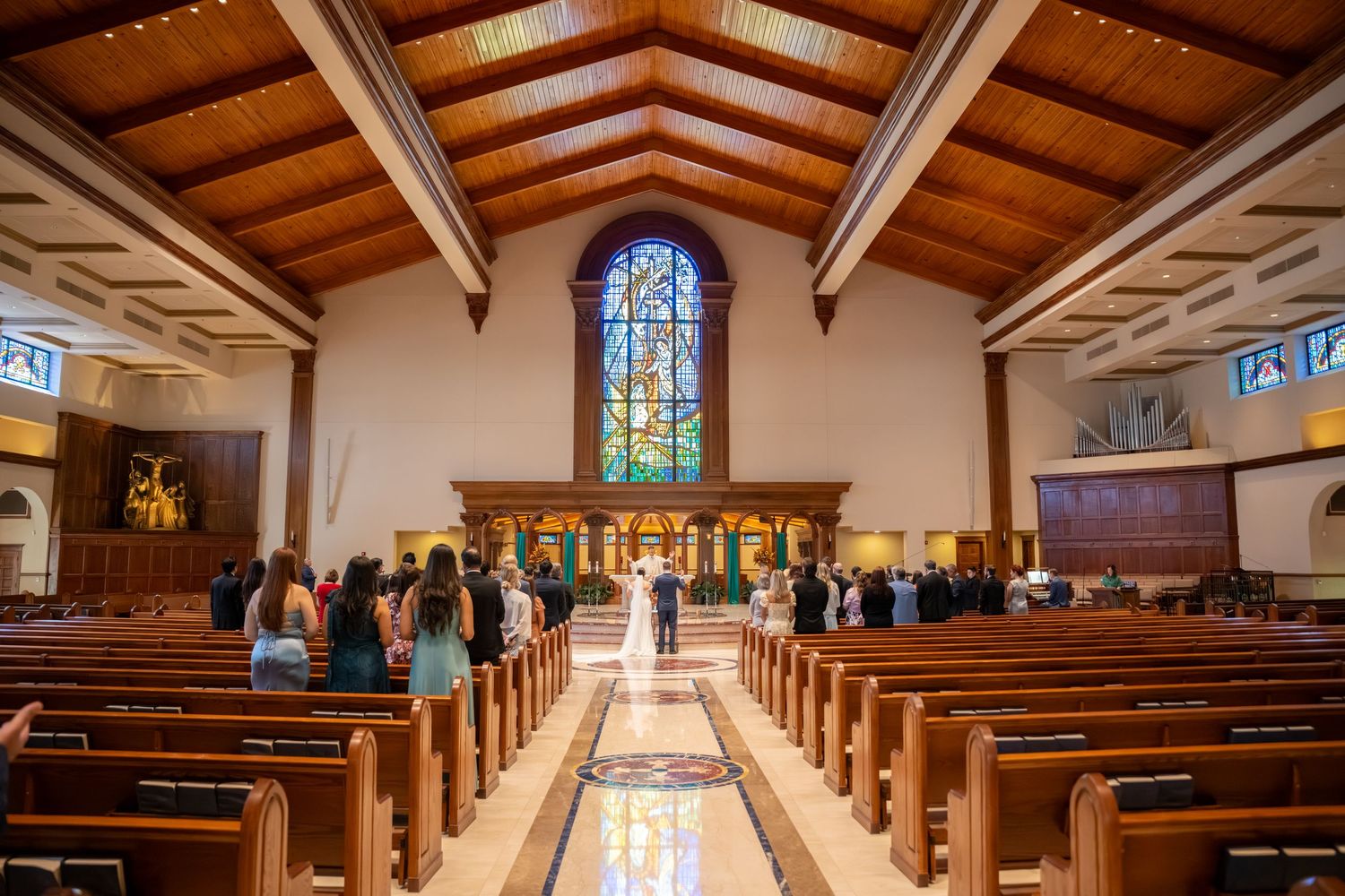 Elegant church wedding ceremony with wooden pews, stained glass window, and guests gathered at the altar.