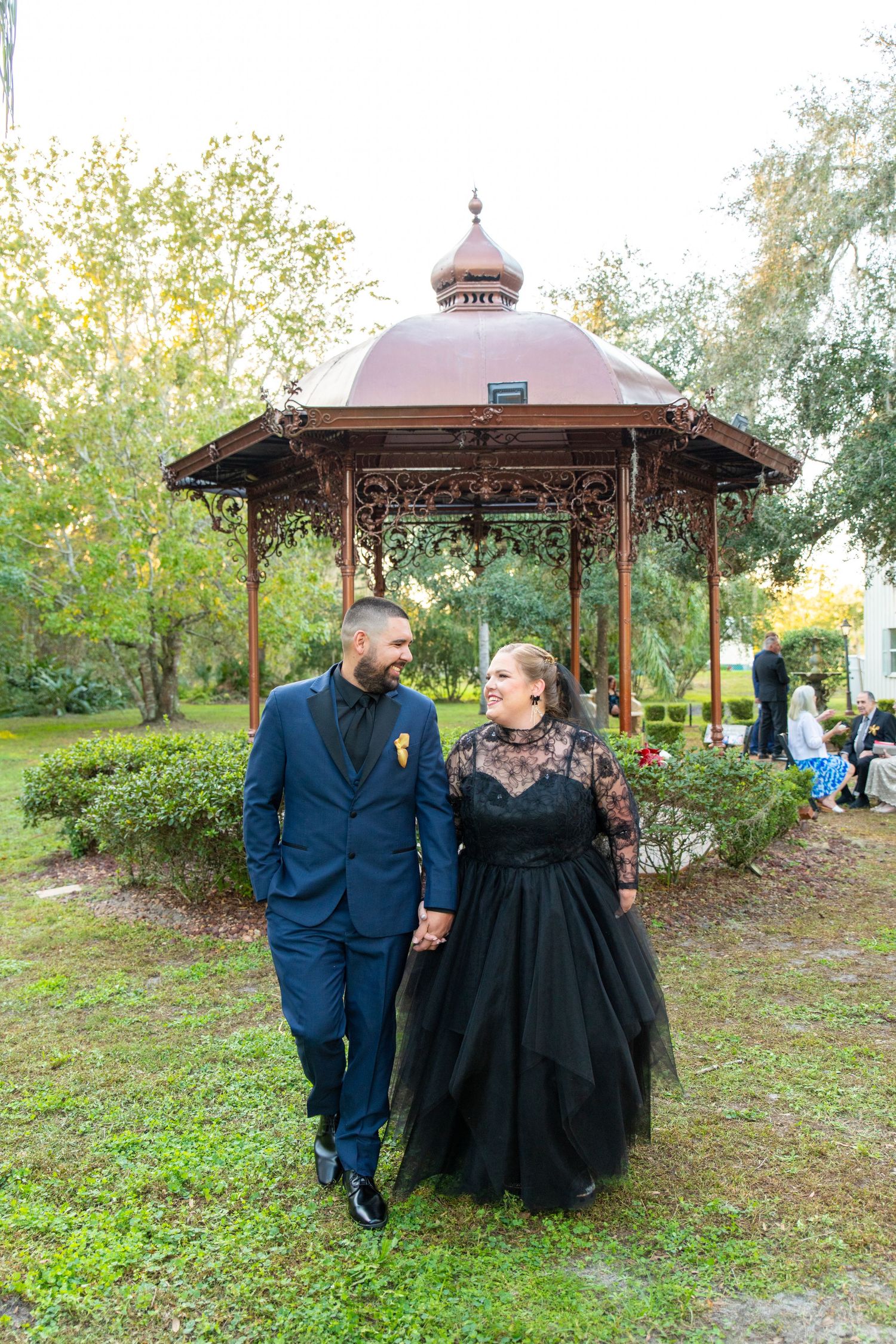 Wedding couple in black outfits walk hand in hand near ornate gazebo in park.