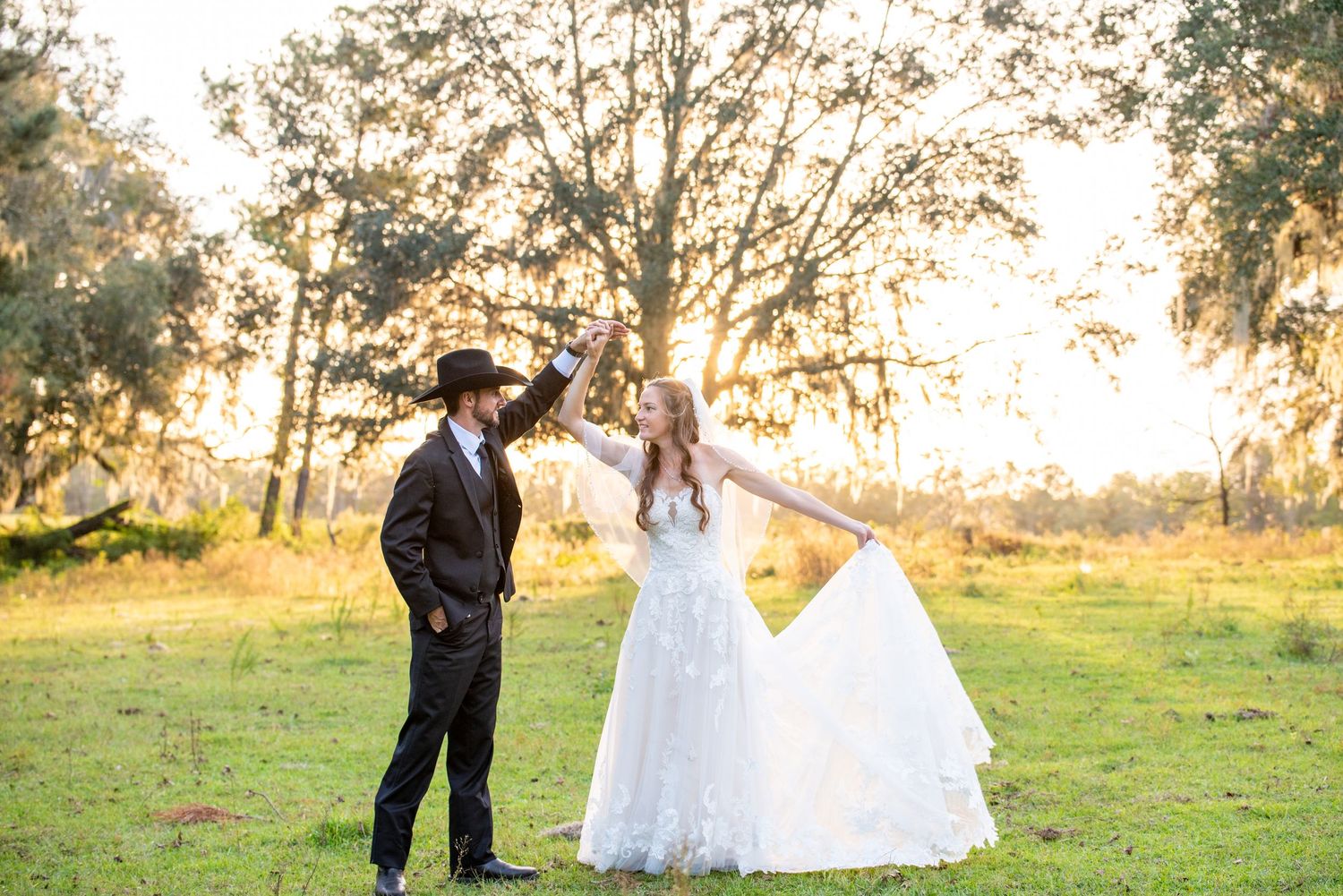 Bride in white gown and groom in black suit dancing outdoors at golden hour in a lush green field.
