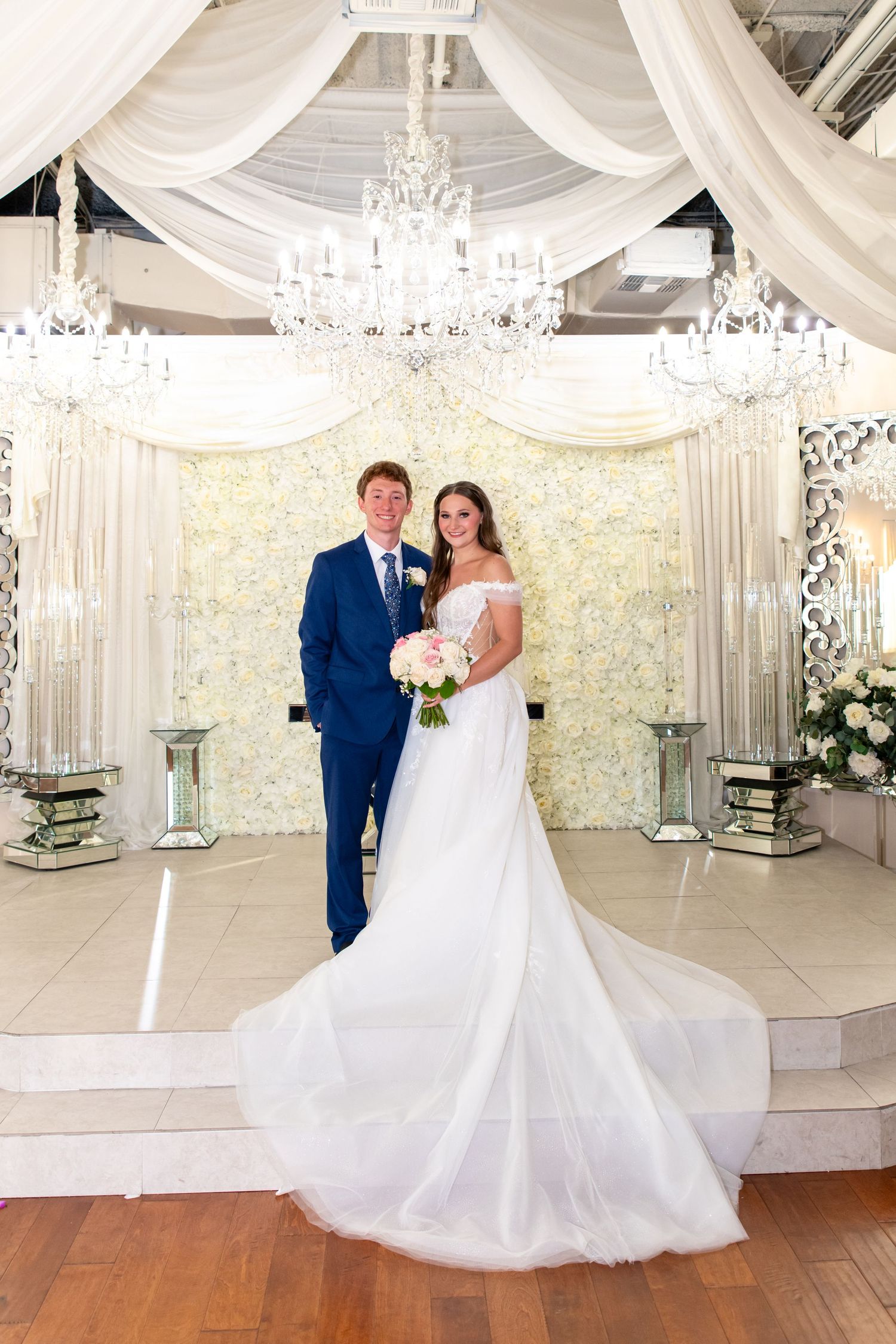 Bride in off-shoulder gown and groom in blue suit pose under crystal chandeliers in elegant ballroom.