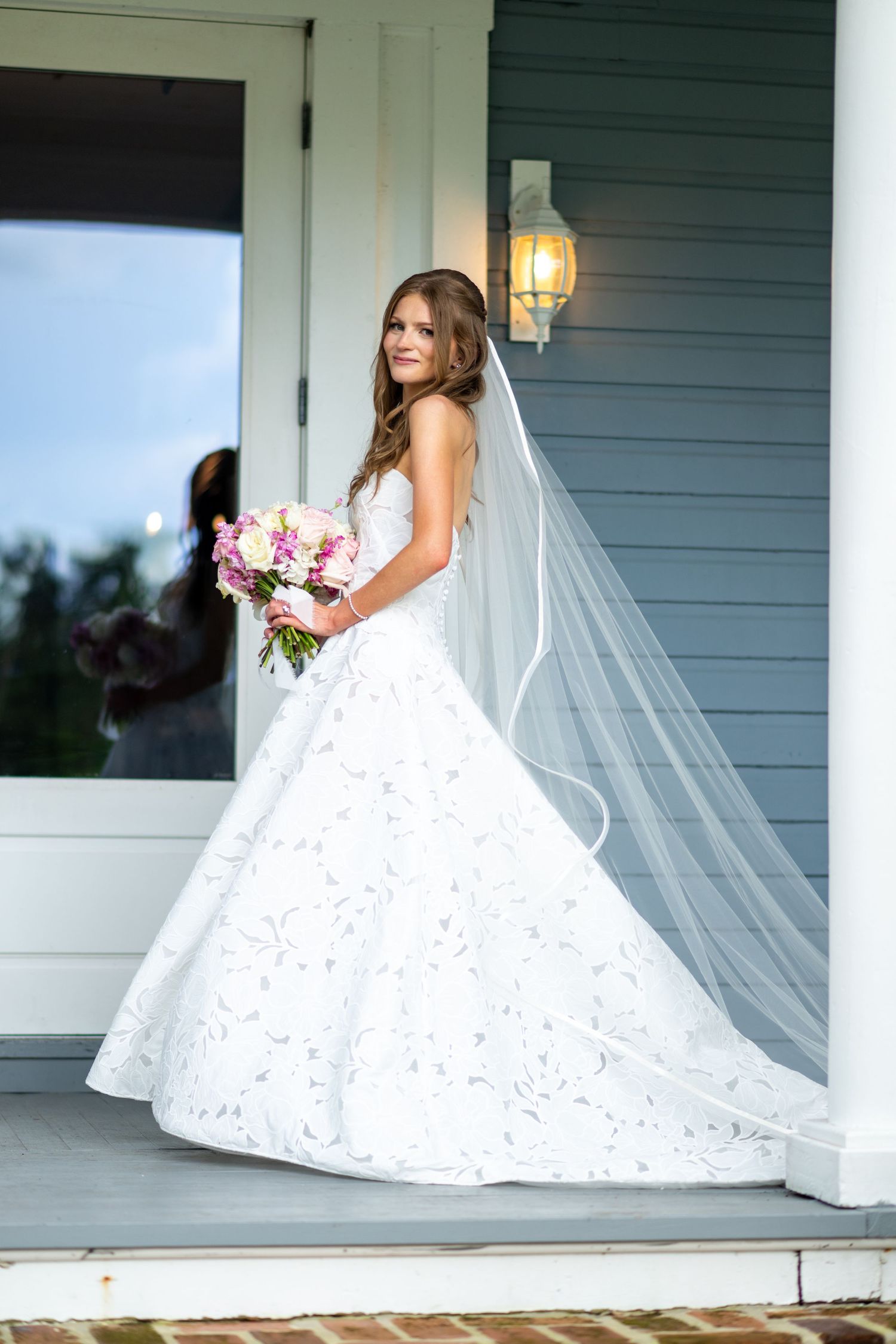 Bride in white ballgown with veil holds pink bouquet on porch steps.