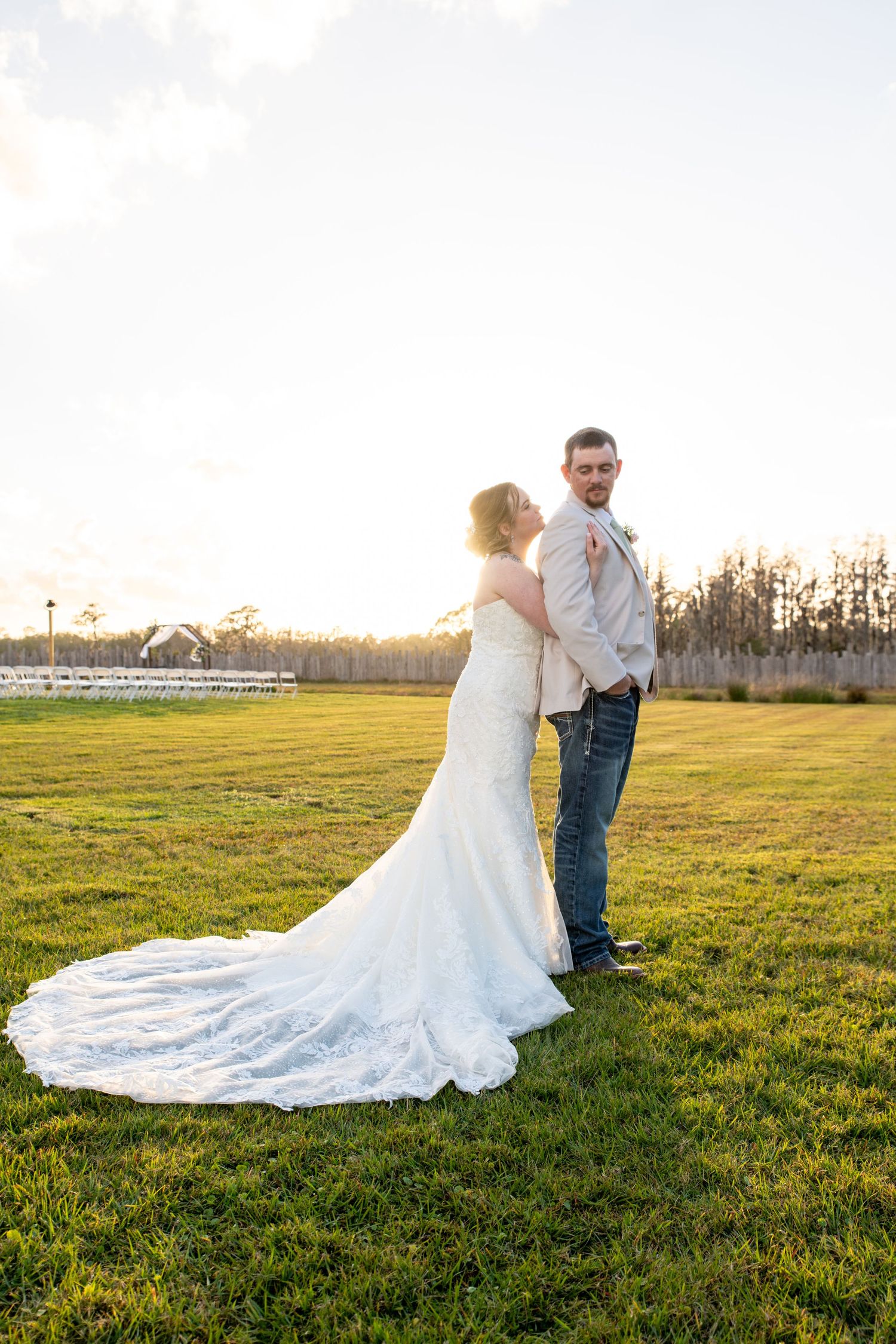Bride in lace gown with long train stands with groom in jeans at golden hour on green field.