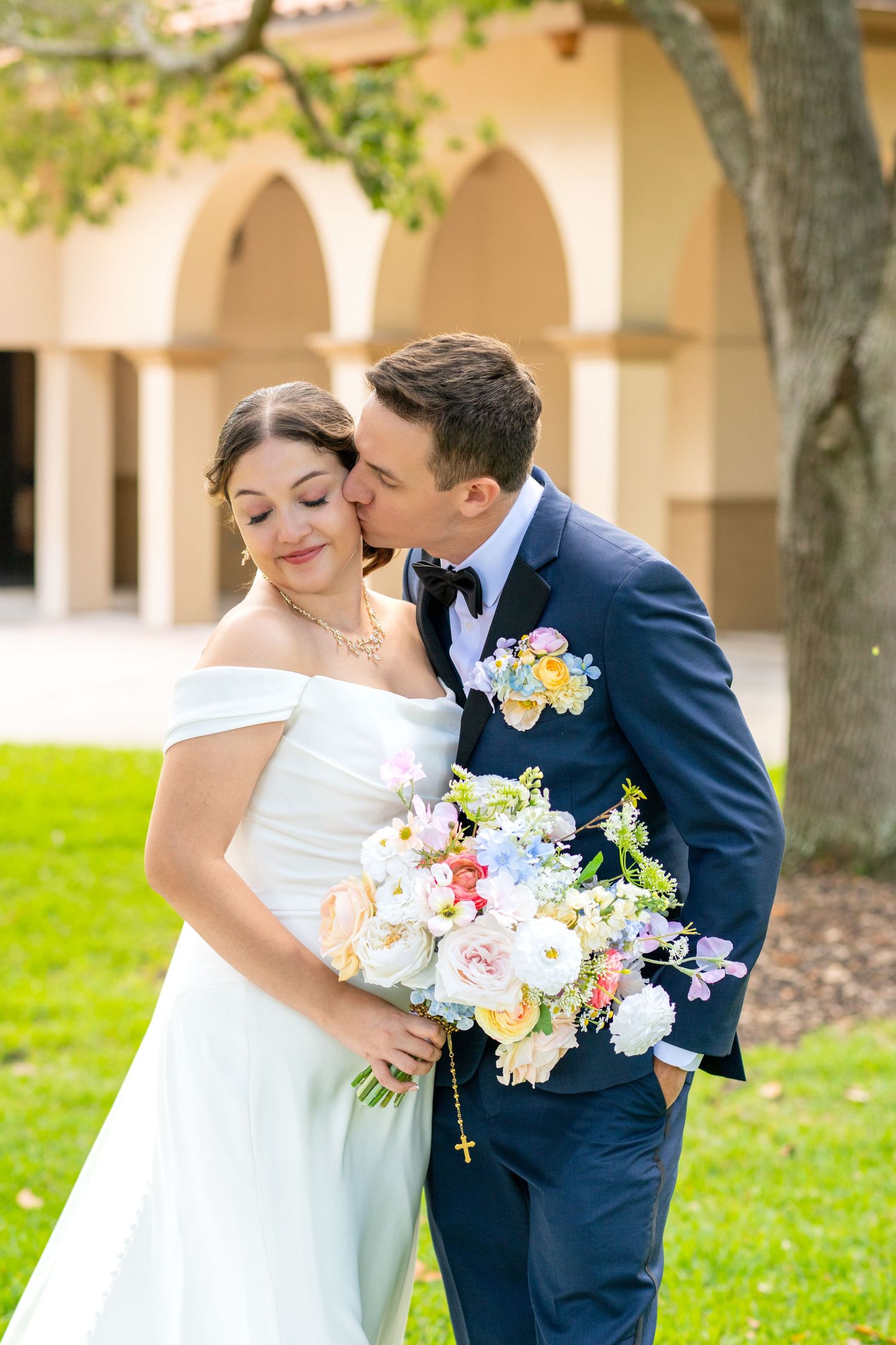 Groom in navy suit kisses bride holding colorful bouquet outside an arched building on wedding day.