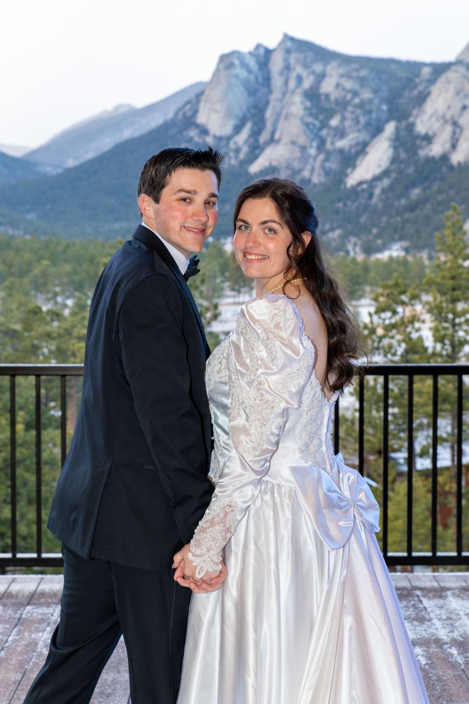 A bride and groom pose together on a mountain balcony, with stunning rocky peaks and pine trees in the background.