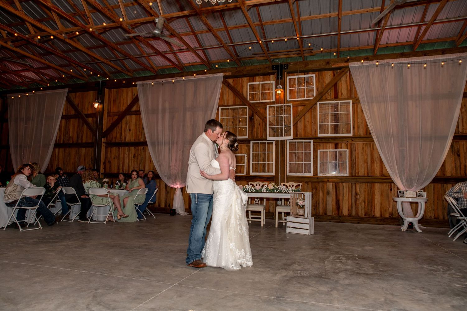 Bride and groom share their first dance in a rustic barn venue decorated with string lights and drapes.