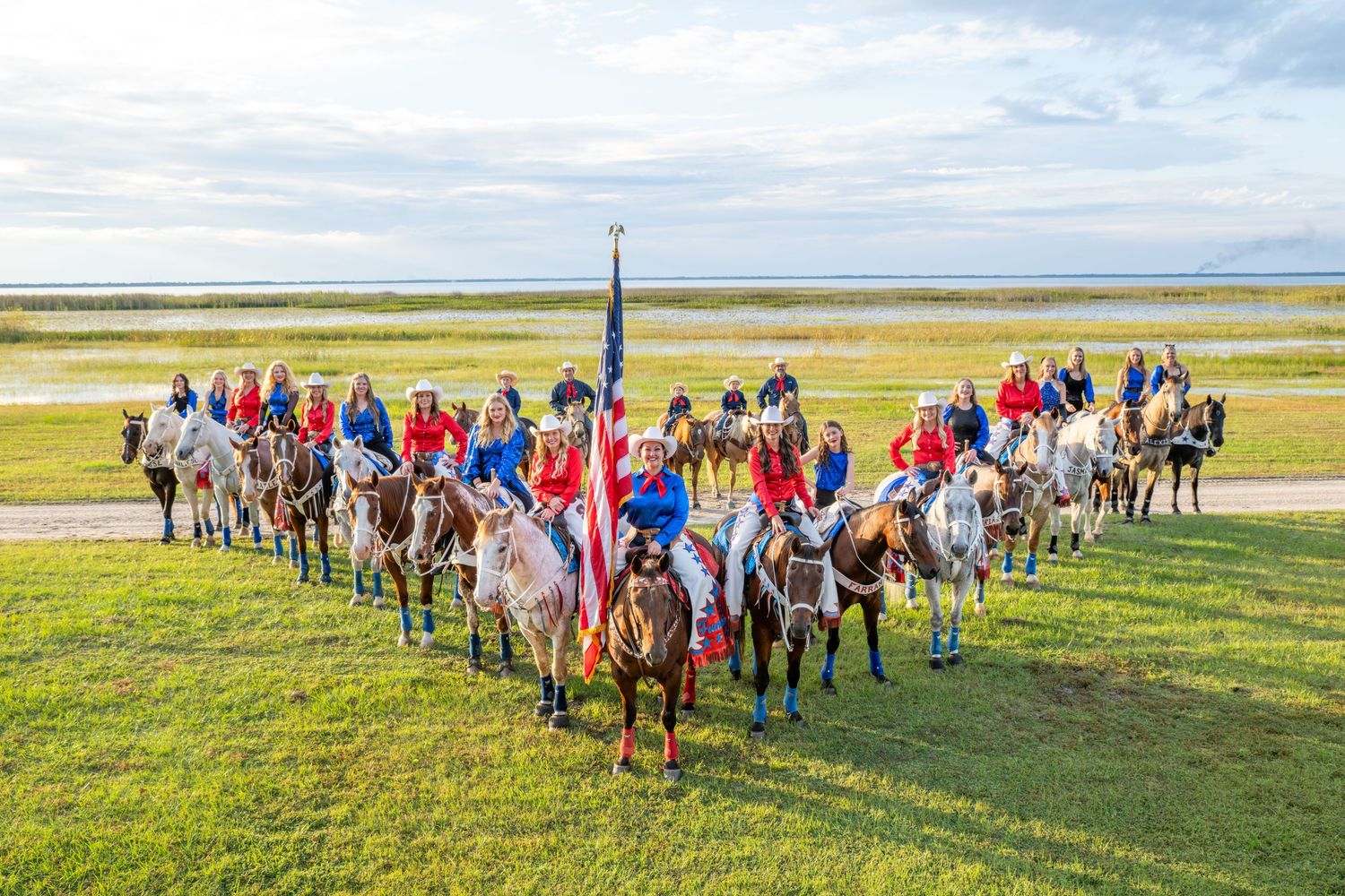 Large group of horseback riders gather outdoors near a waterfront carrying an American flag for a patriotic equestrian event.