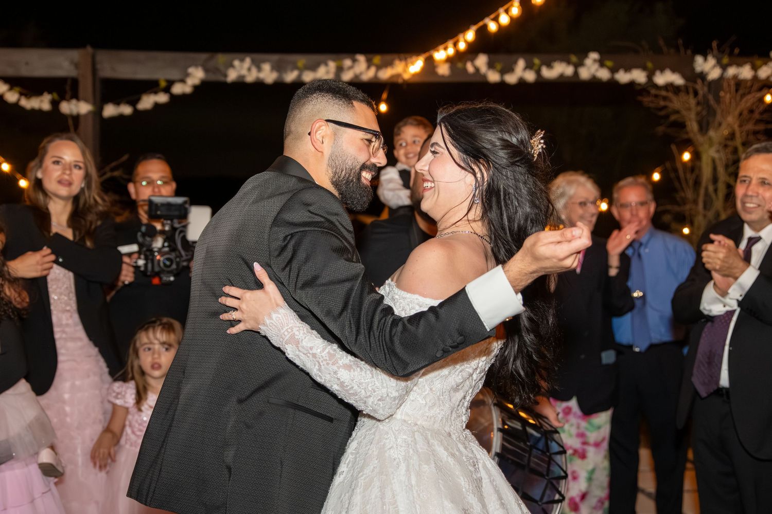 Bride and groom embrace warmly during a lively outdoor wedding reception at night.