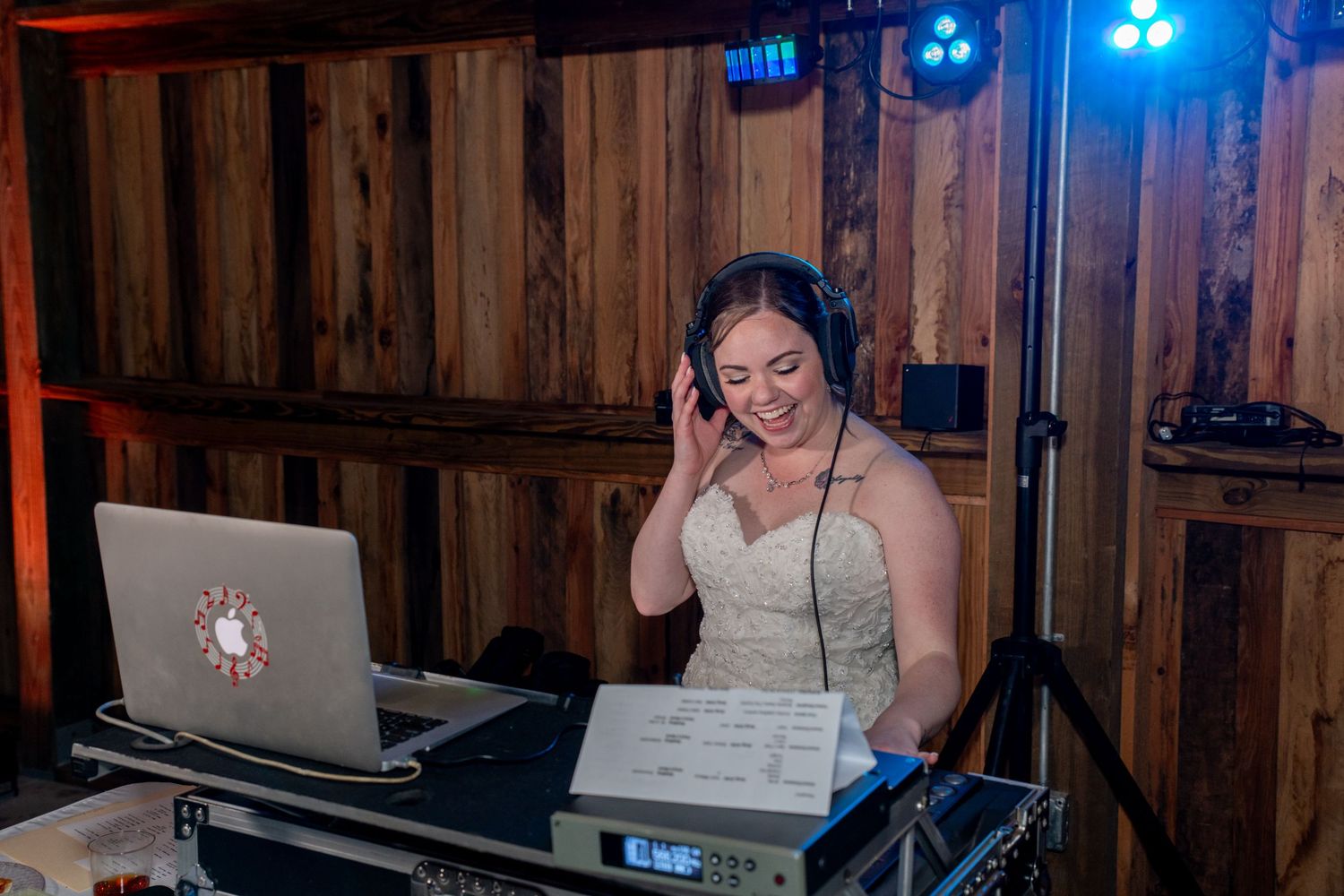 Smiling female DJ wearing headphones performs at a wedding reception in a rustic barn venue with DJ equipment.