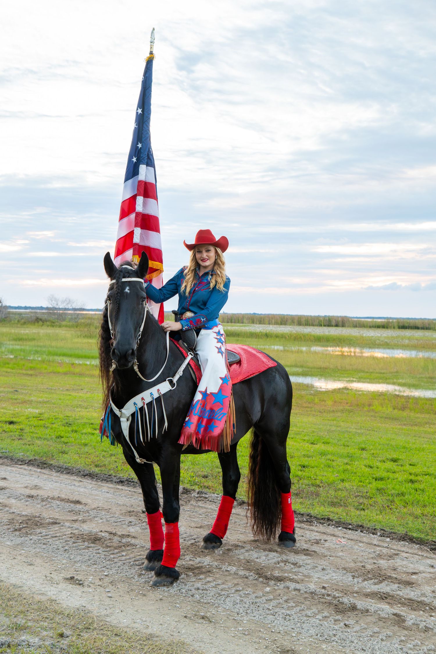 Blonde cowgirl in red hat rides black horse while holding American flag on rural dirt path.