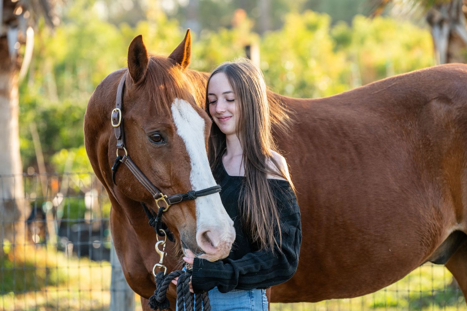 Young woman with long hair affectionately bonding with a chestnut horse wearing a halter outdoors.
