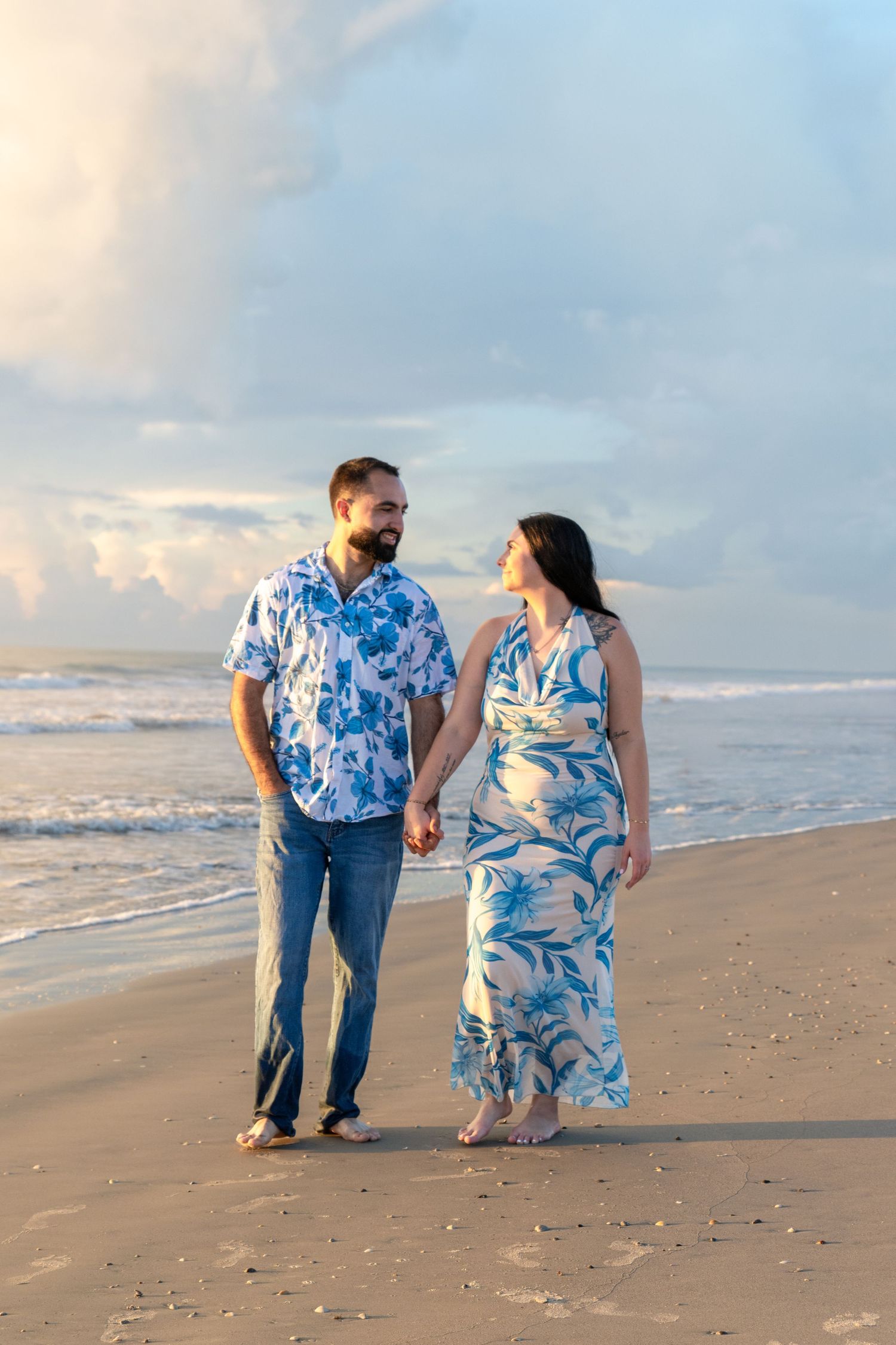 Couple in matching blue floral outfits holding hands and walking on a beach at sunset.