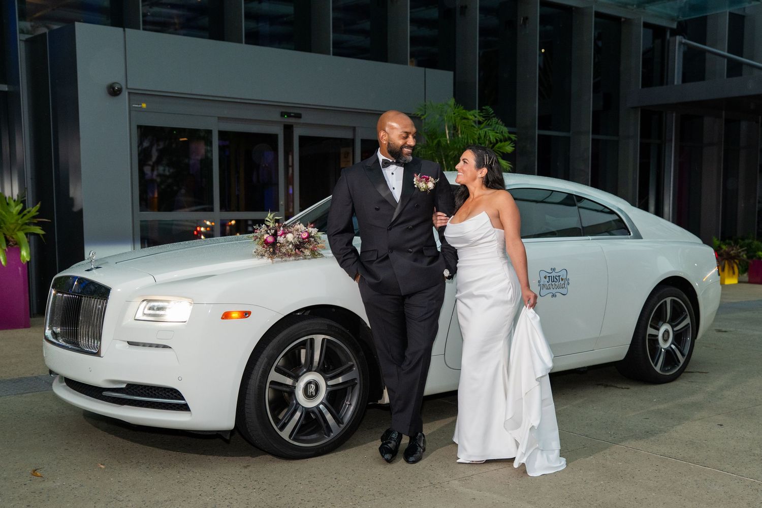 Elegant wedding couple posing beside a white Rolls-Royce, bride in white gown and groom in black tuxedo.