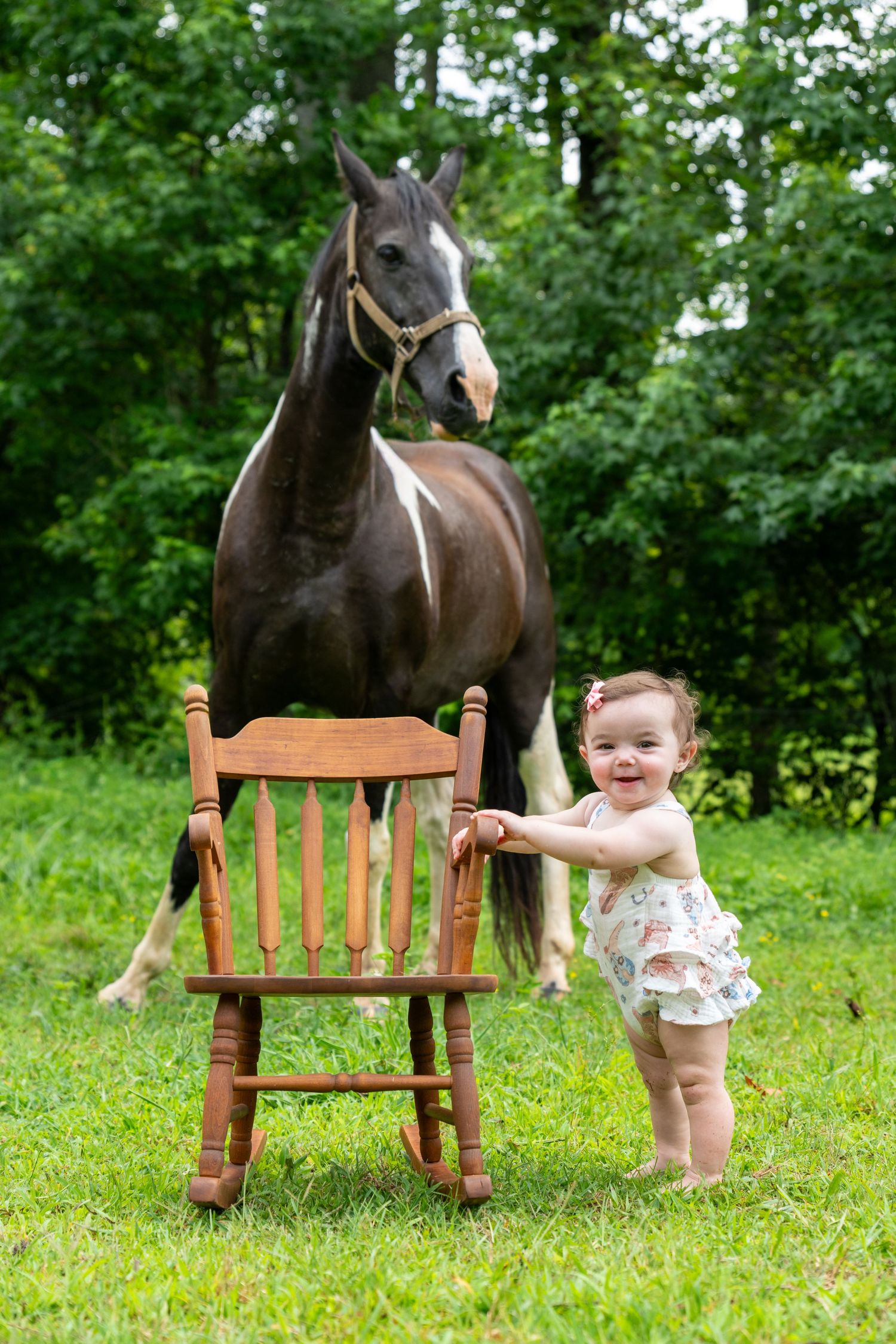 Smiling baby girl standing by wooden chair with dark horse on green grass outdoors.