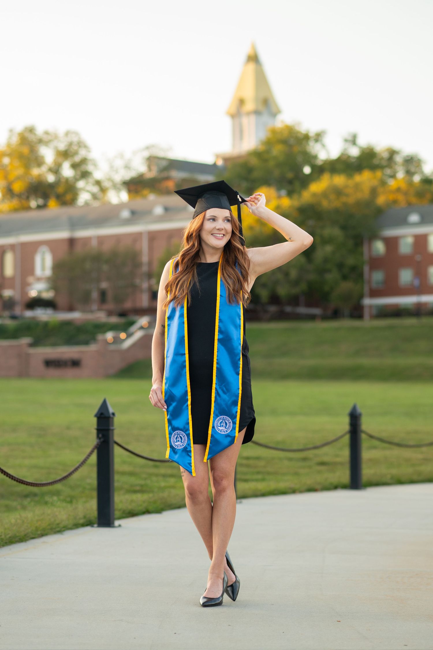 Smiling graduate in cap and blue stole walks on campus path with historic building in background.