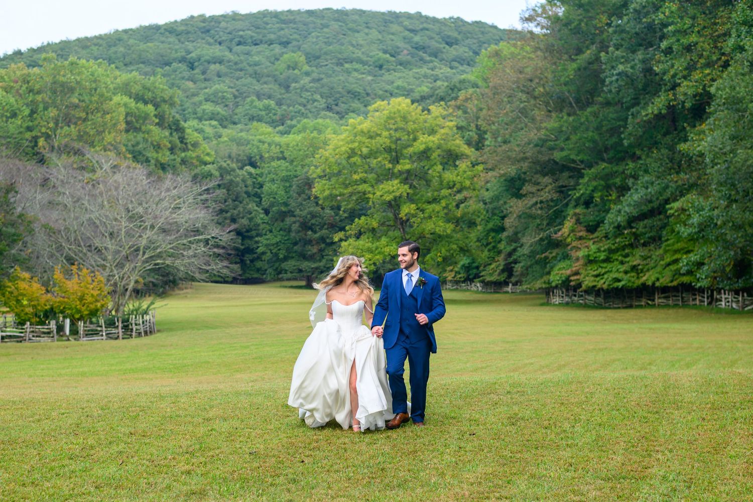 Bride and groom walking hand in hand across lush green lawn with wooded hills in background on wedding day.