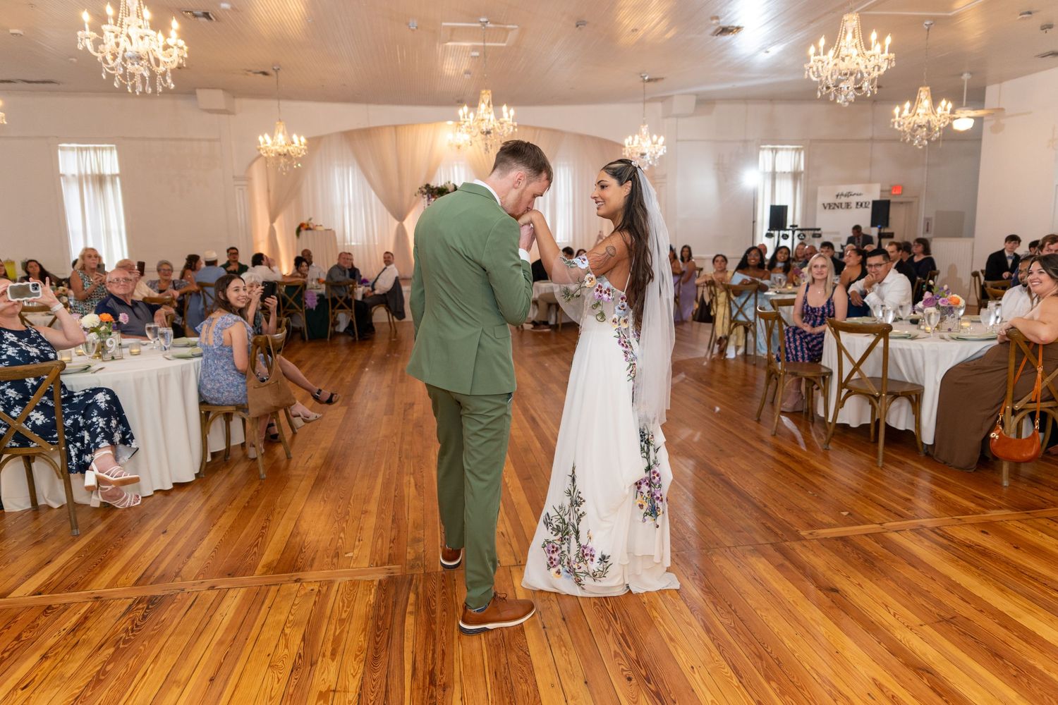 Bride and groom share first dance on wood floor in elegant ballroom with chandeliers and wedding guests seated at tables.