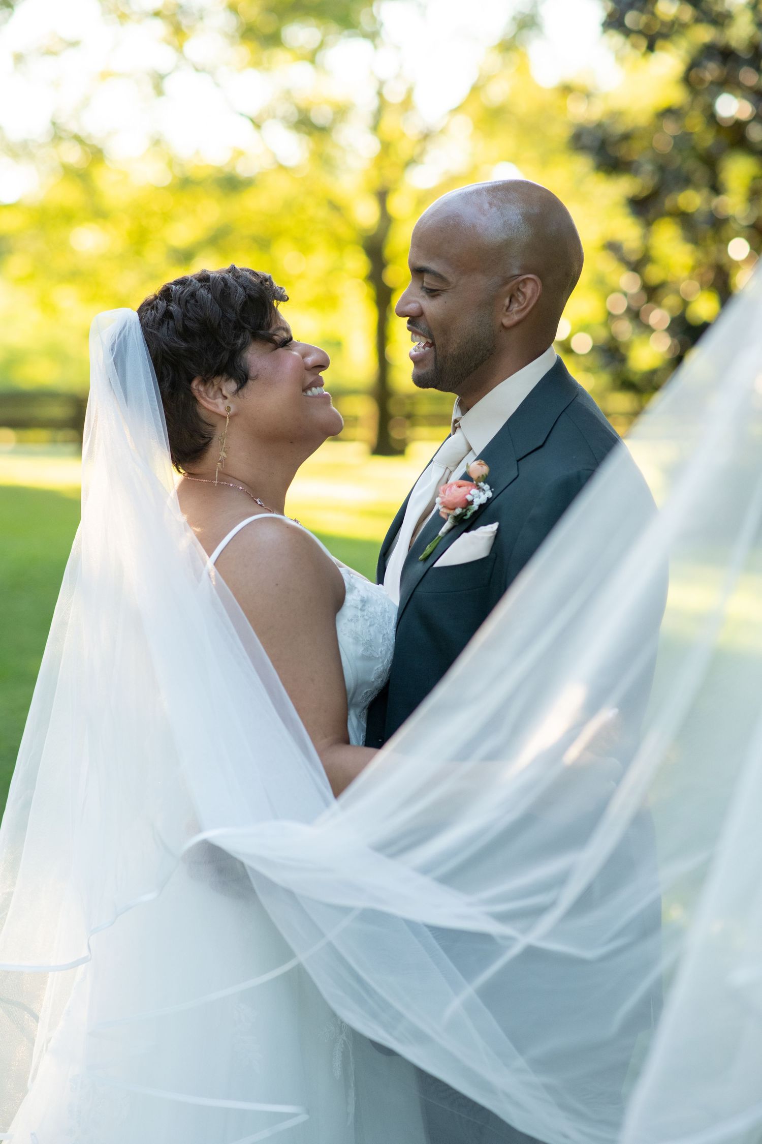 Smiling bride and bald groom embrace outdoors with flowing veil in foreground.