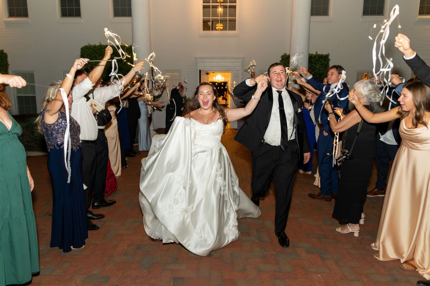 Bride and groom exit wedding reception through cheering guests waving streamers at elegant venue.