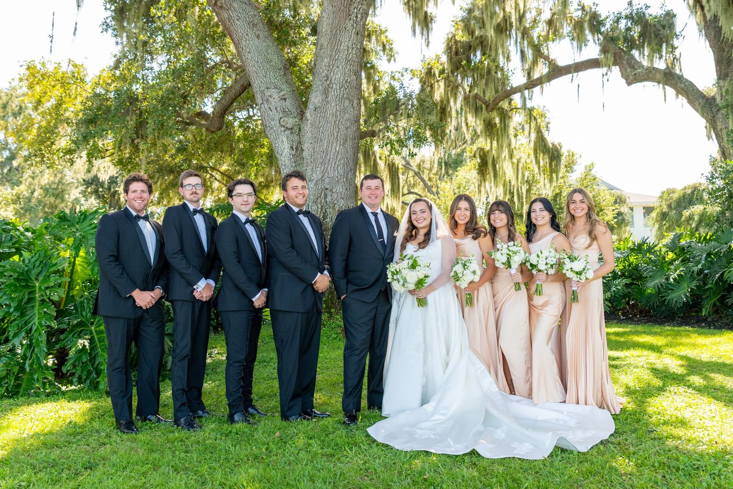 Wedding party posing outdoors under a large oak tree, groomsmen in black tuxedos and bridesmaids in champagne gowns.