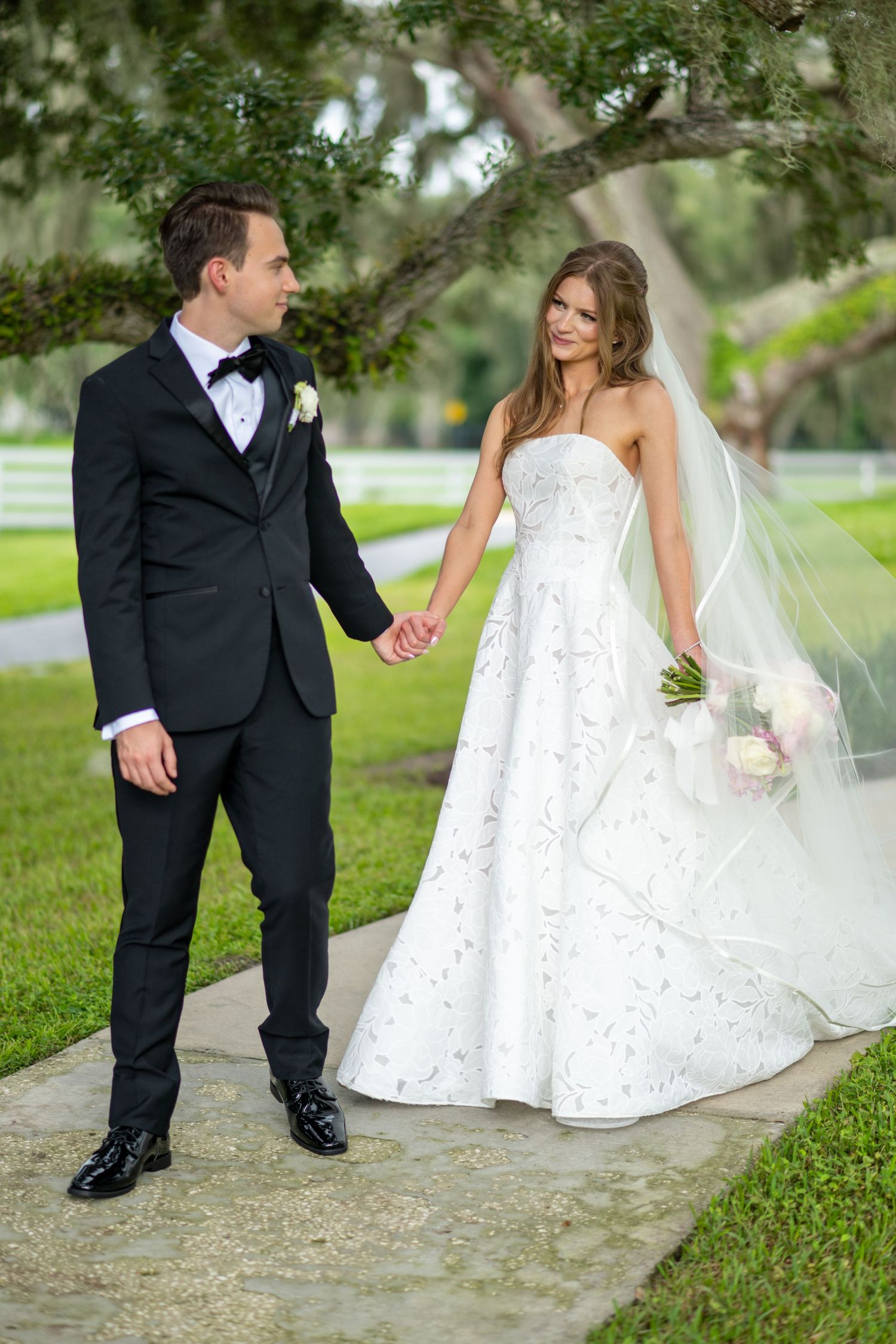Bride and groom holding hands walking on a garden path, she in a strapless gown with veil, he in a black tuxedo.