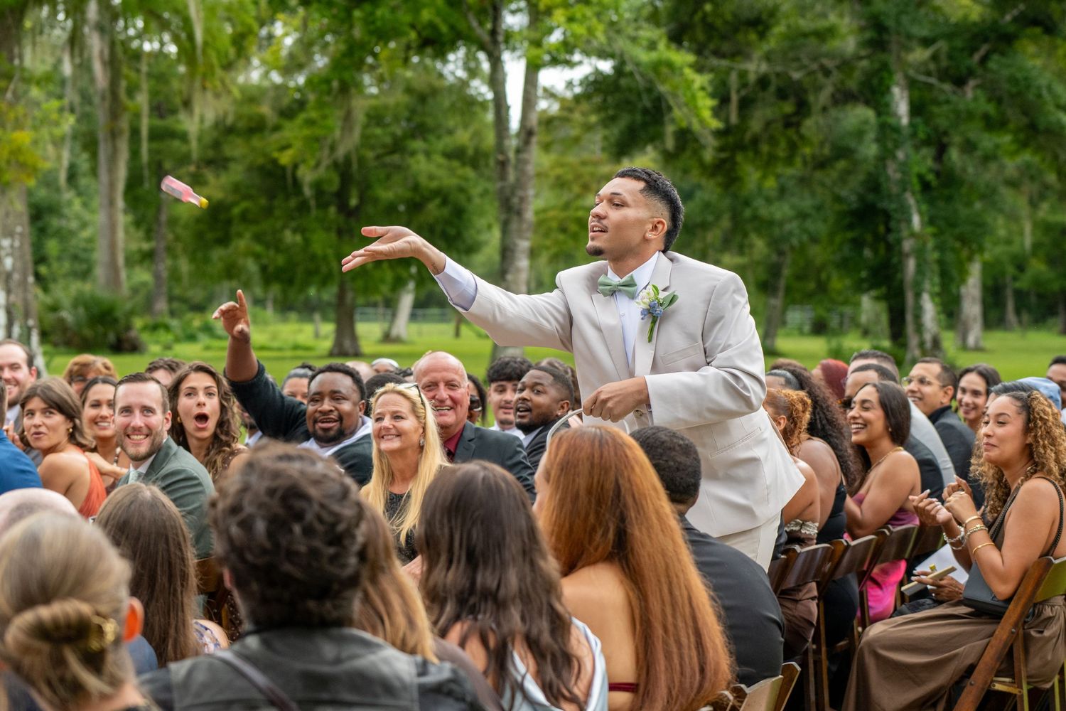 Groom in gray suit tosses bouquet to cheering wedding guests seated outdoors amid lush green trees.