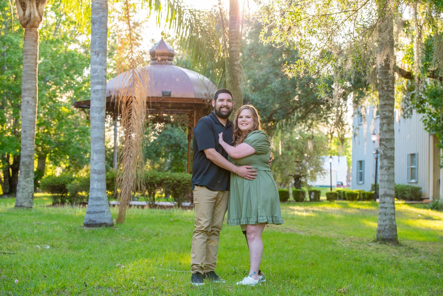 Smiling couple embracing in a lush green park with palm trees and a gazebo behind them.