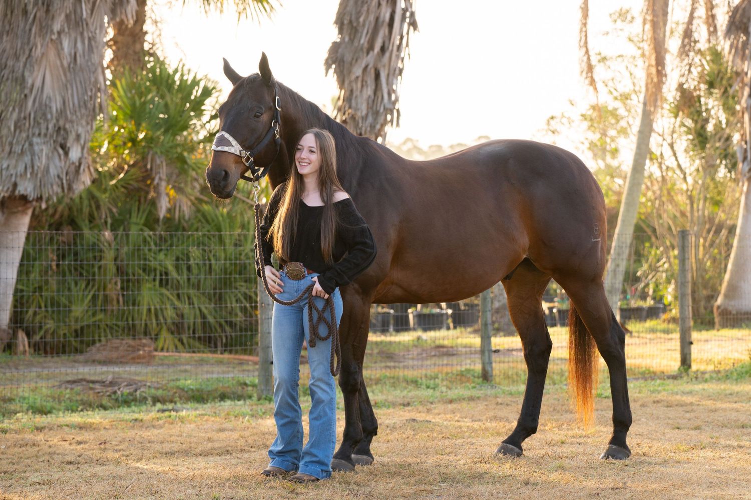 Teen girl standing beside a tall dark bay horse at golden hour on a ranch.
