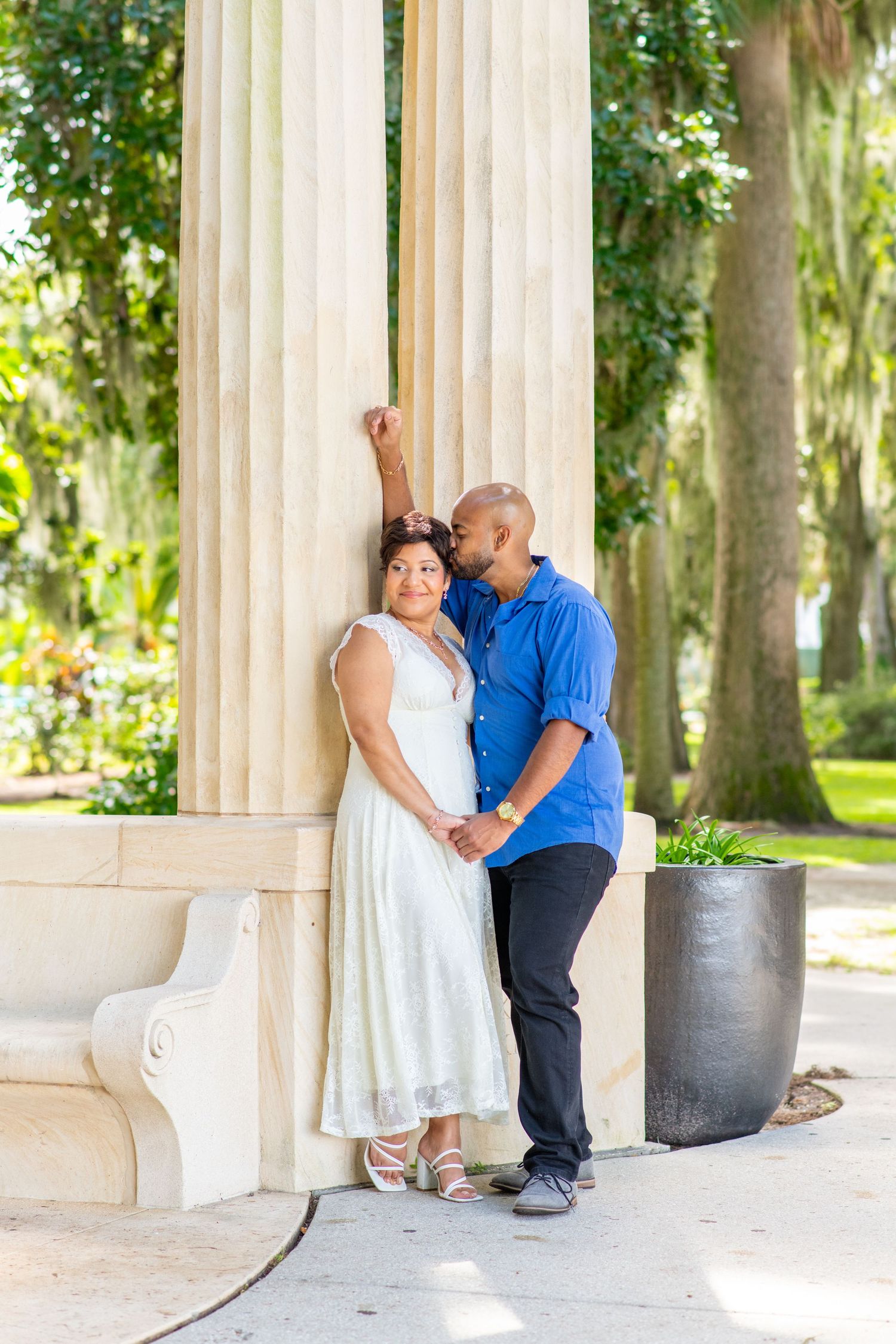 Couple posing by marble columns in a lush park, woman in white dress, man in blue shirt leaning close.