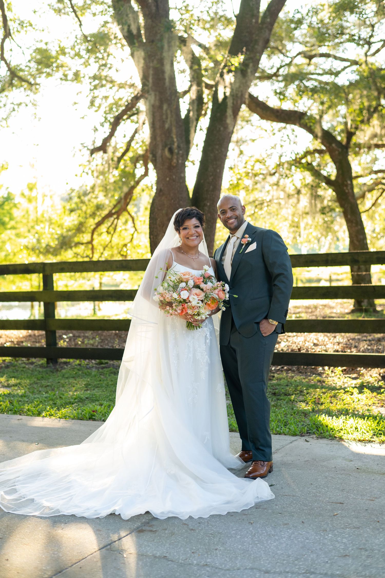 Bride and groom pose together outdoors in wedding attire, she holds a peach bouquet, large oak tree and fence behind them.