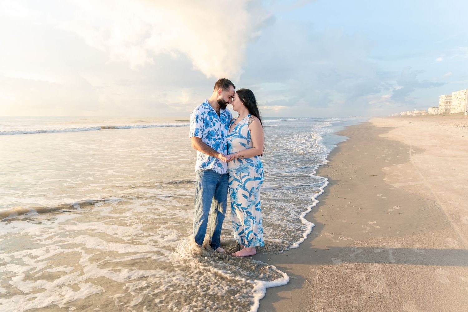 Couple embracing on a sandy beach at sunset, waves washing over their feet, wearing blue outfits.