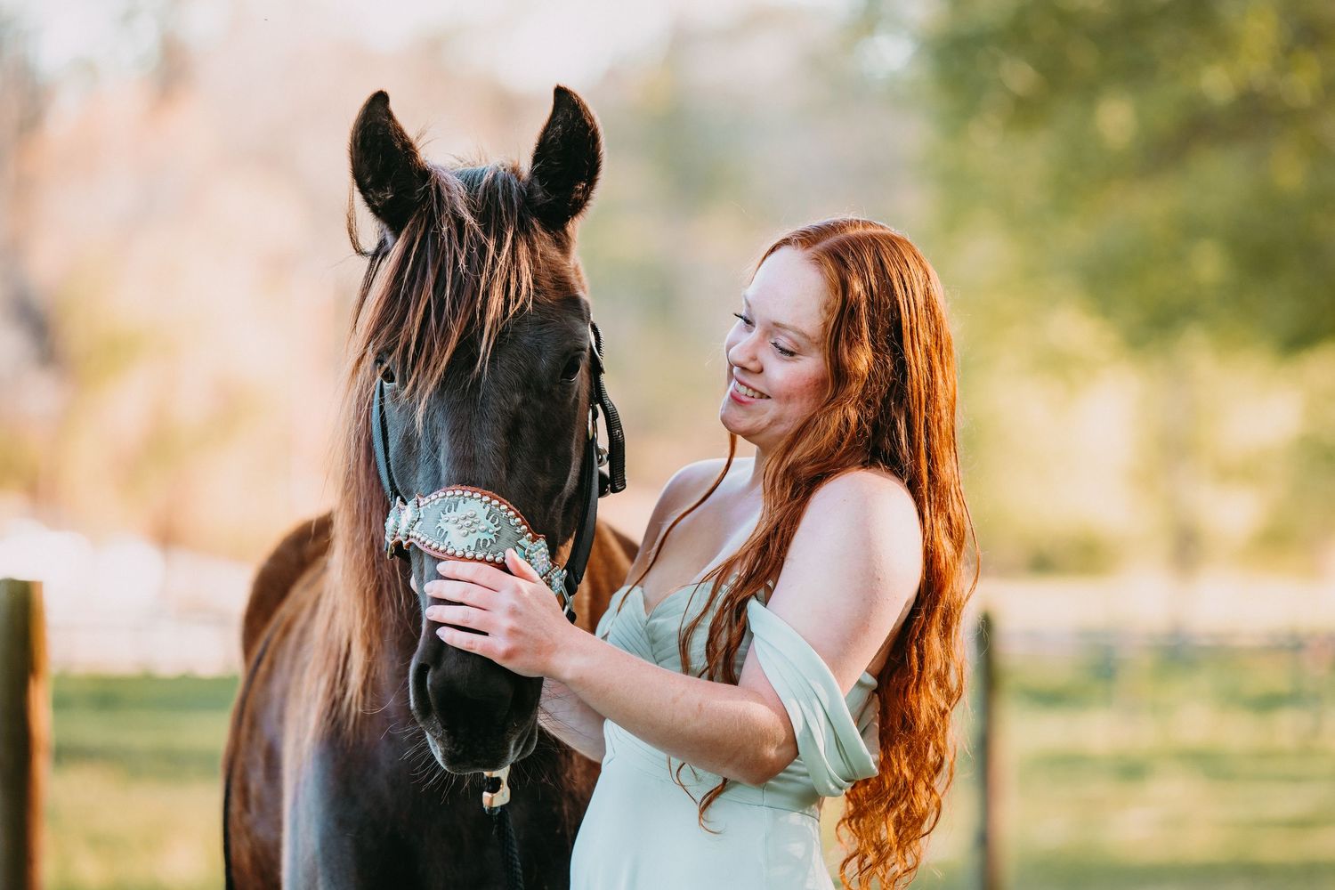 Red-haired woman in mint dress tenderly holding a dark horse's face outdoors in golden light.