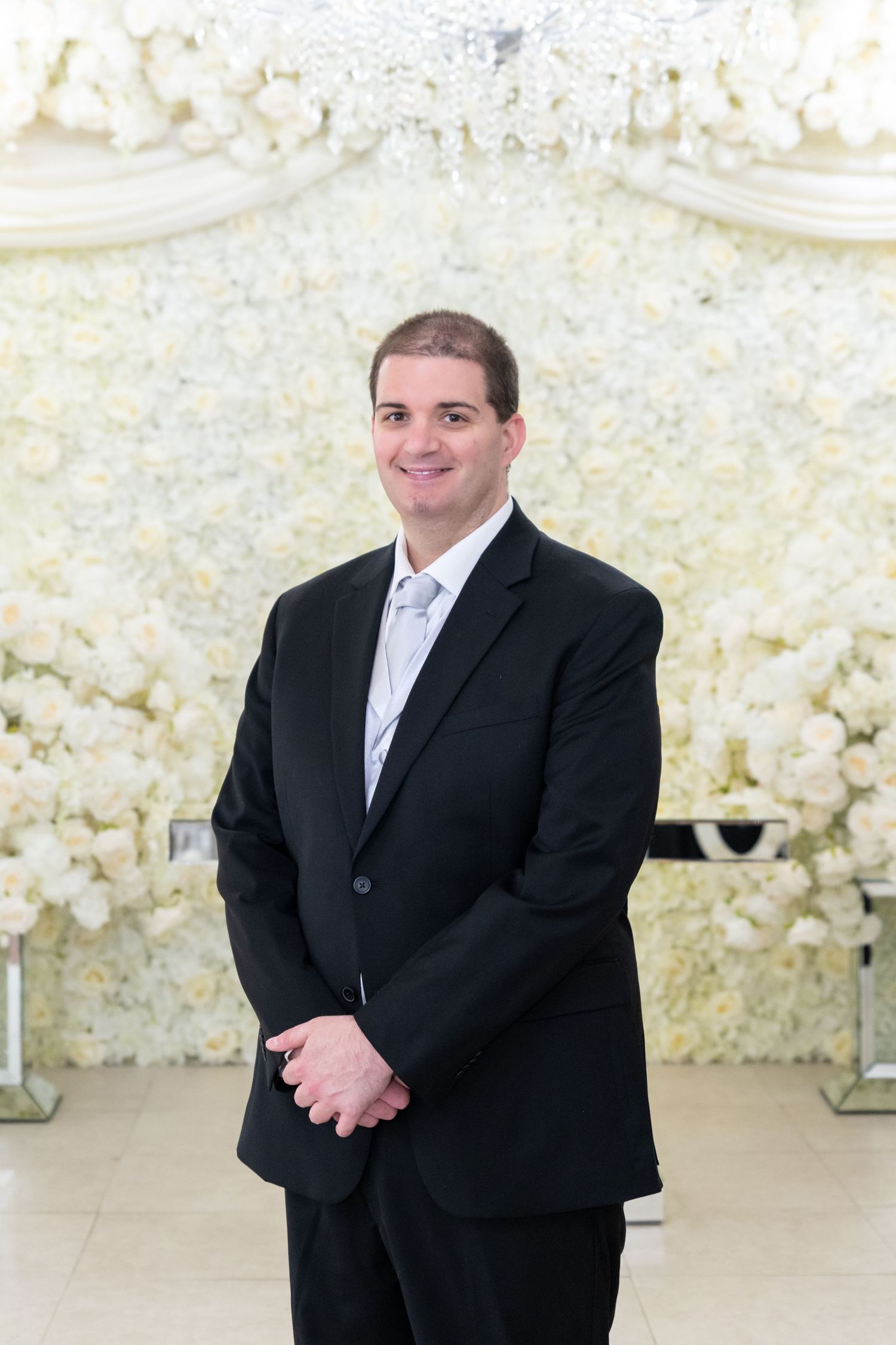 A smiling man in a black suit and silver tie stands before an elegant white floral wall backdrop at a wedding venue.