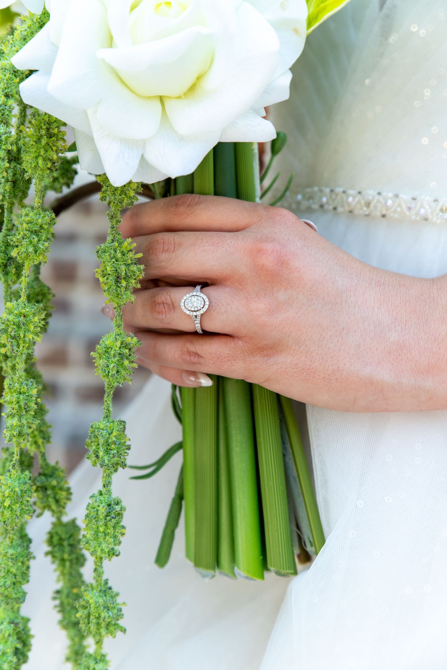 Bride holding white floral bouquet with green stems, wearing oval diamond halo engagement ring on wedding day.