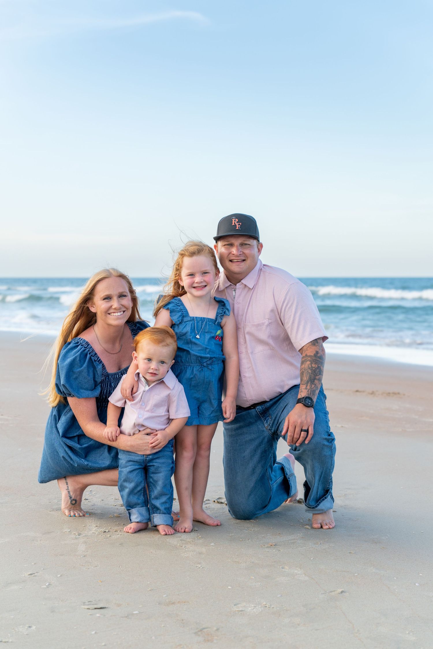 Happy family of four posing together on a sandy beach with ocean waves in the background.