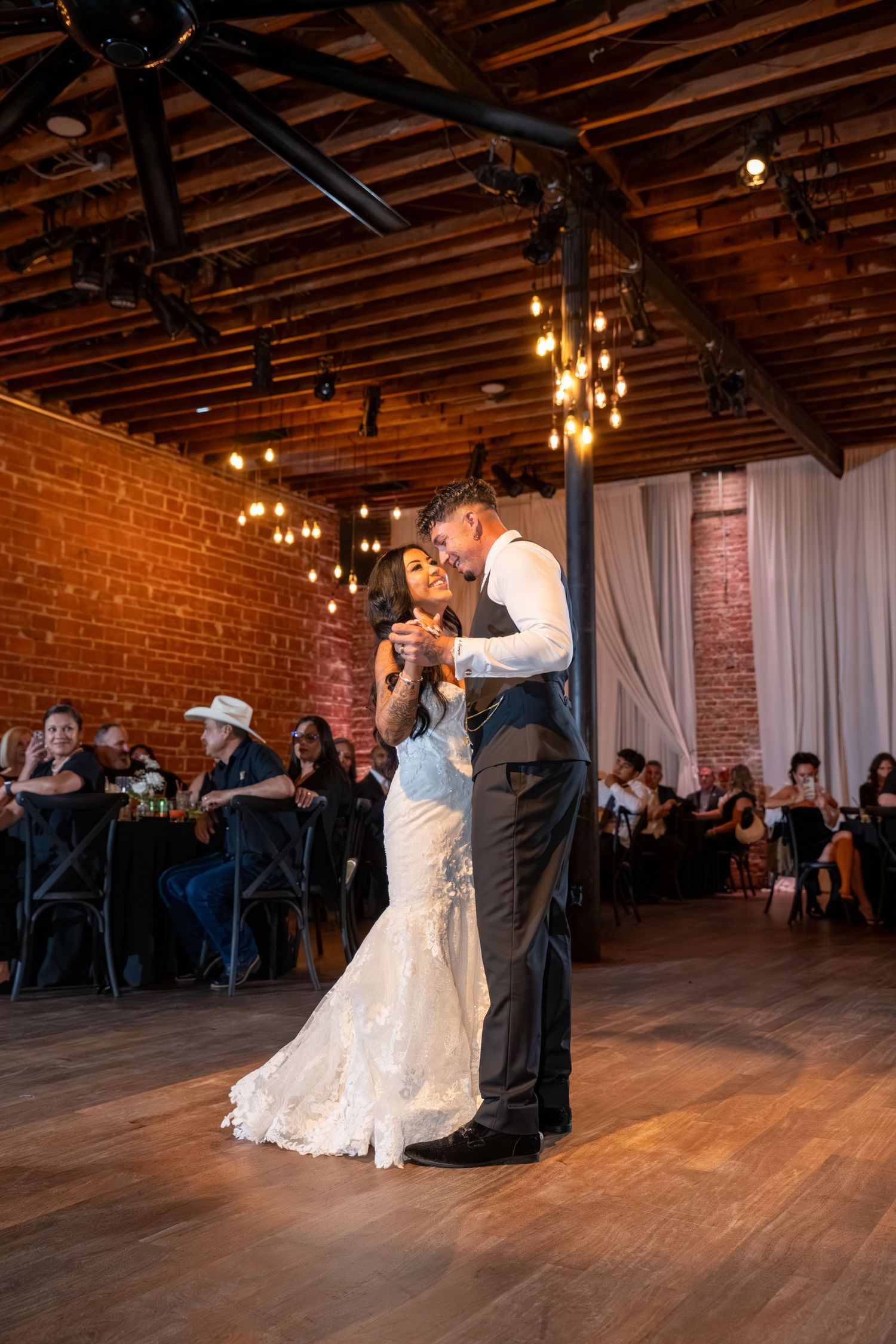 Bride and groom share their first dance in a rustic brick venue with Edison bulb lighting and guests seated around the dance floor.