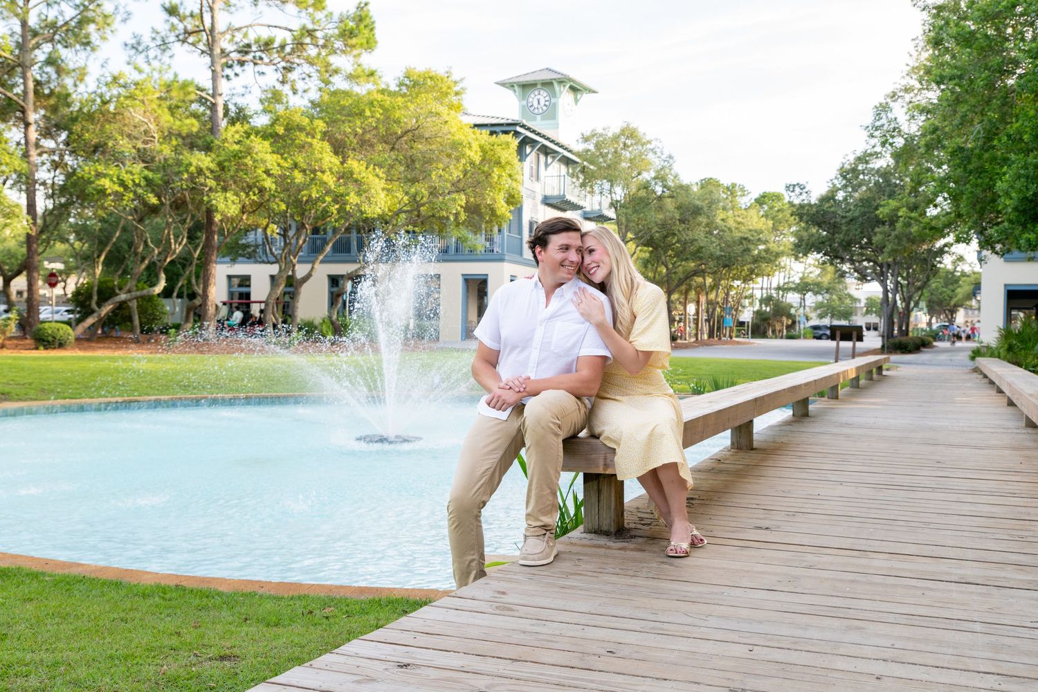 Couple sitting on a wooden boardwalk by a fountain, surrounded by lush trees and a building in the background.