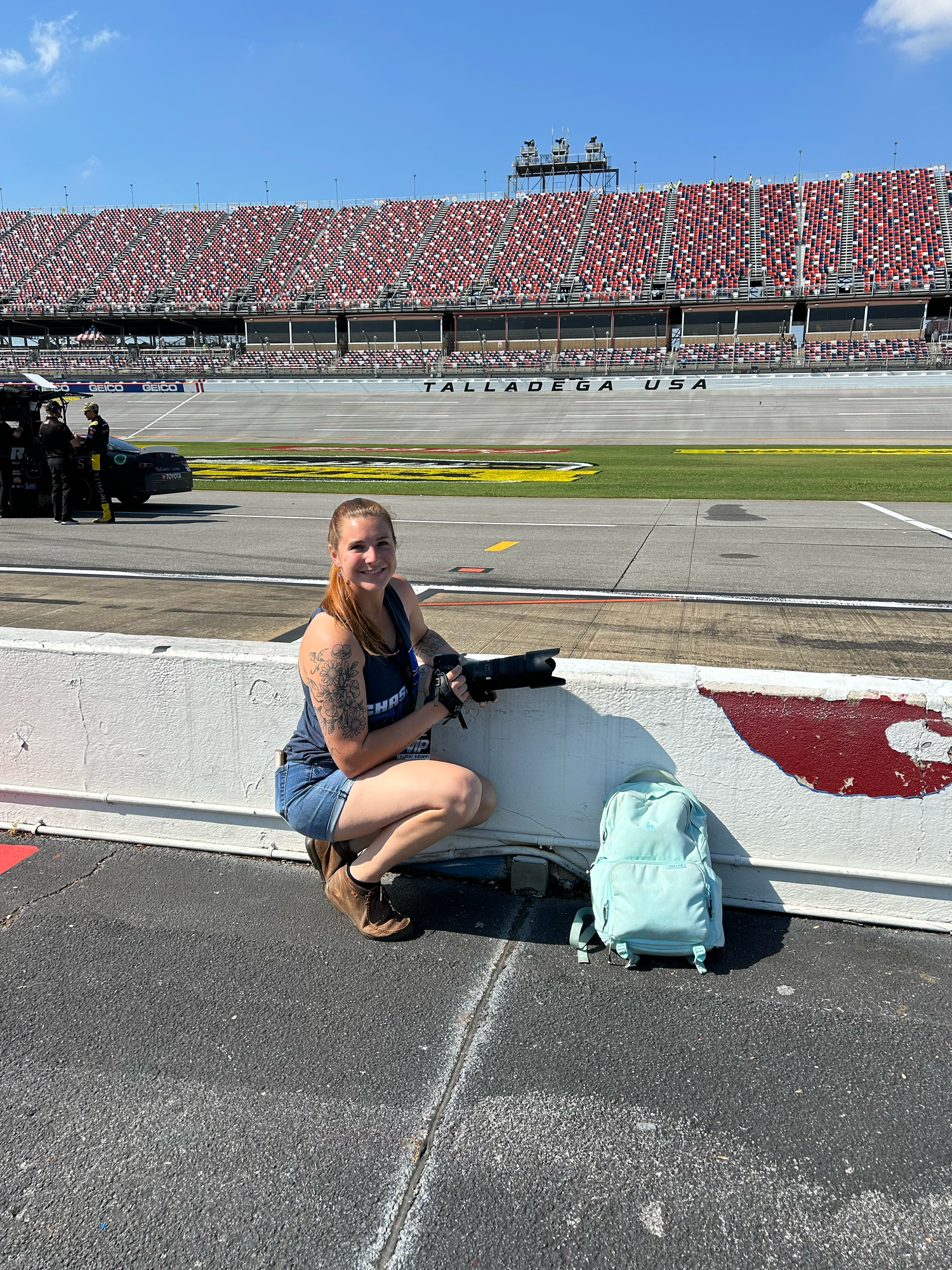 Woman posing at Talladega USA NASCAR racetrack with a light blue backpack on a sunny day.
