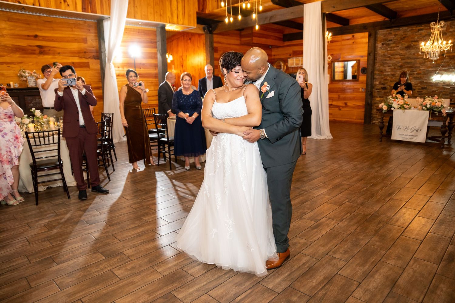 Bride and groom share their first dance on a wooden floor surrounded by guests in a rustic wedding venue.