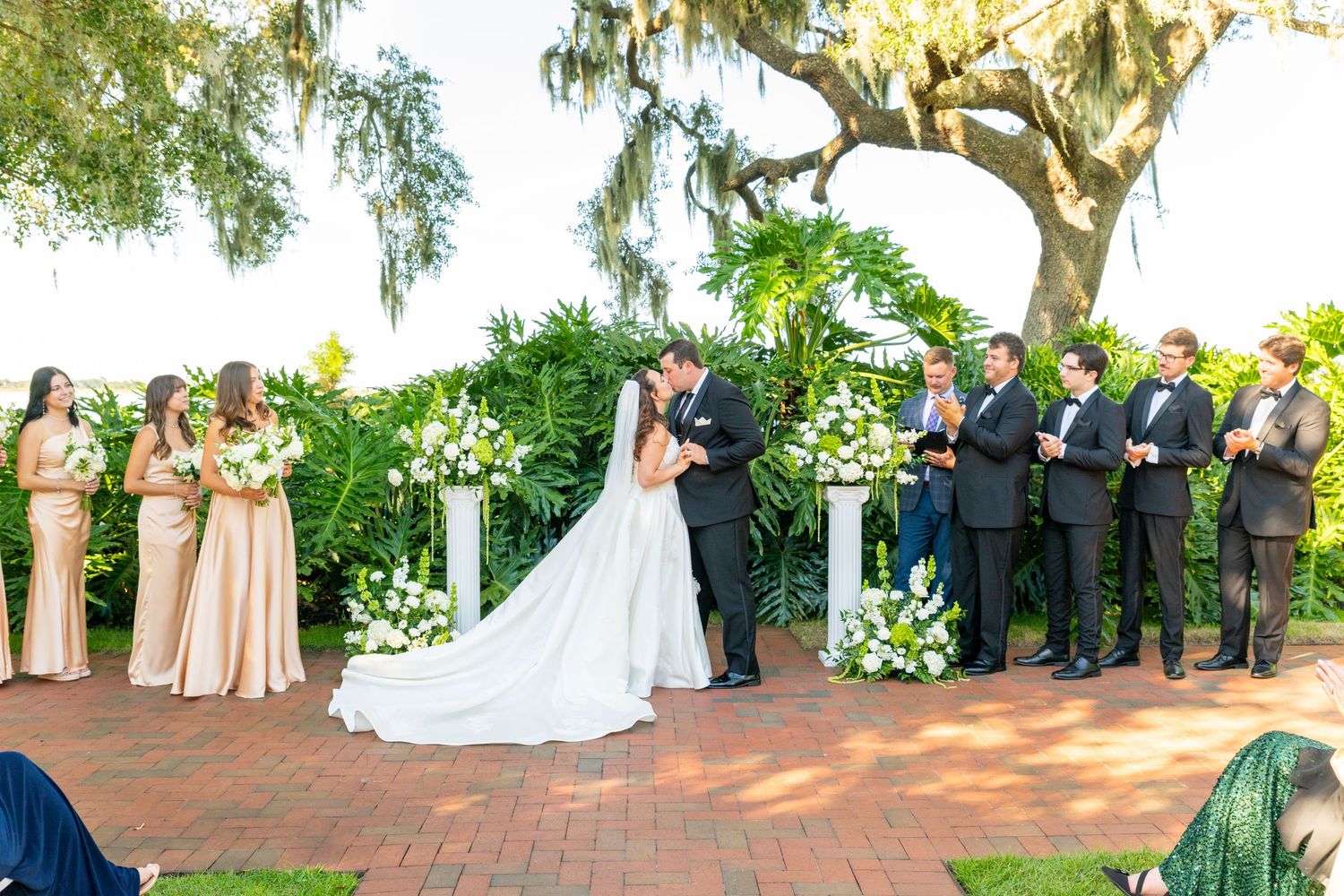 Bride and groom share first kiss at outdoor wedding ceremony with bridesmaids and groomsmen beside lush greenery backdrop.