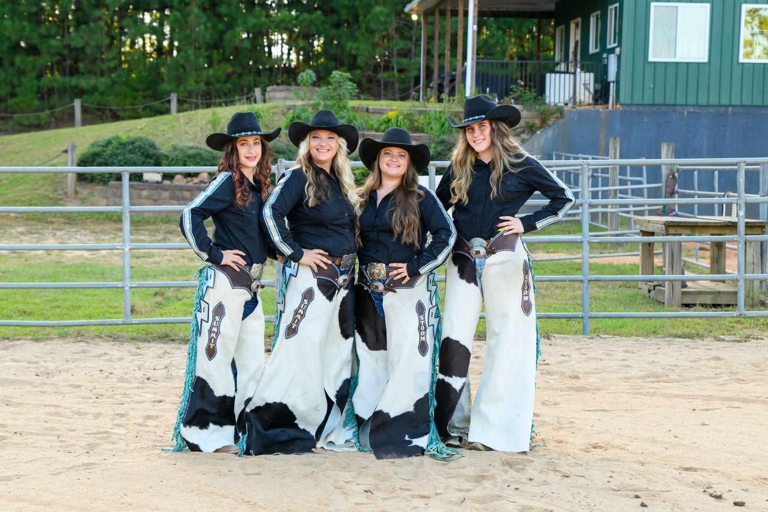 Four women in matching black cowboy hats and cow-print skirts pose together at an outdoor equestrian arena.