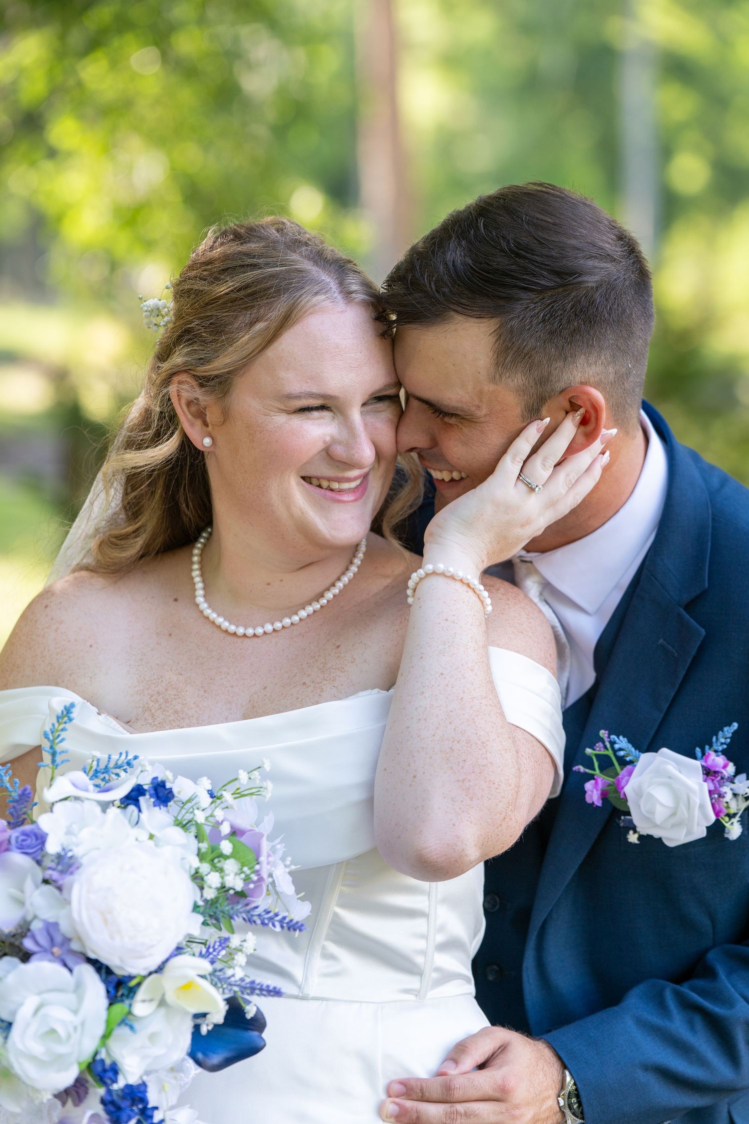 Smiling bride in white off-shoulder gown holds purple bouquet as groom in navy suit nuzzles her cheek outdoors.