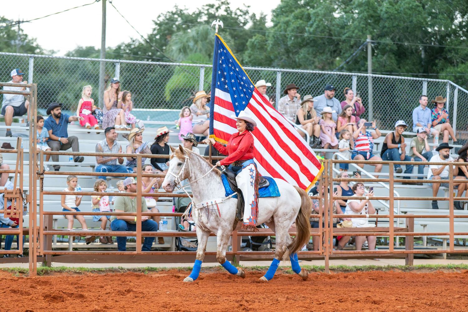 A rider on a white horse carries the American flag at a rodeo arena with spectators in the stands.