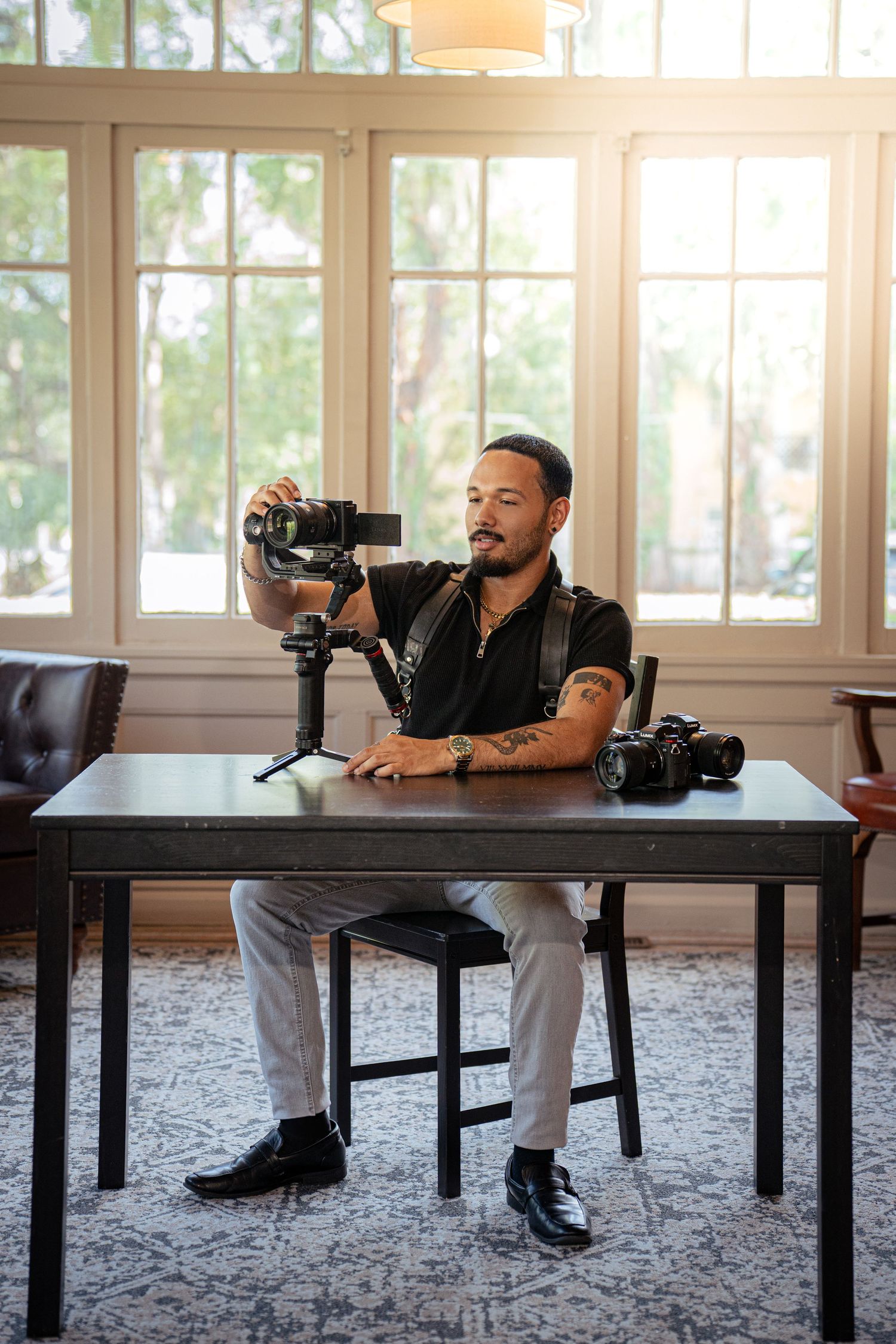 Bearded man in a black shirt sits at a wooden table holding a camera on a gimbal stabilizer, with a second camera beside him.
