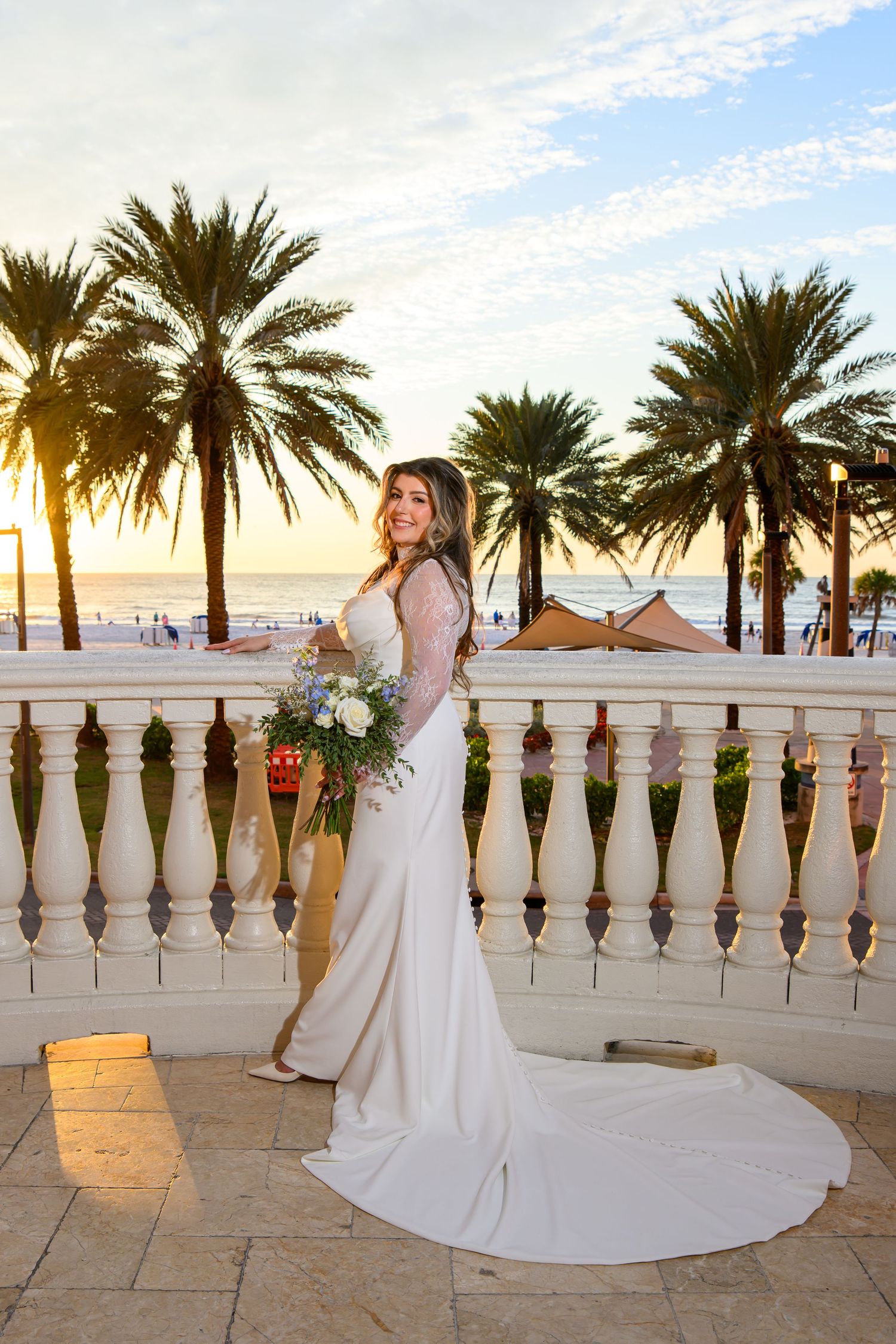 Bride in a long white lace gown holding a blue and white bouquet on a stone balcony with palm trees and a sunset beach view.