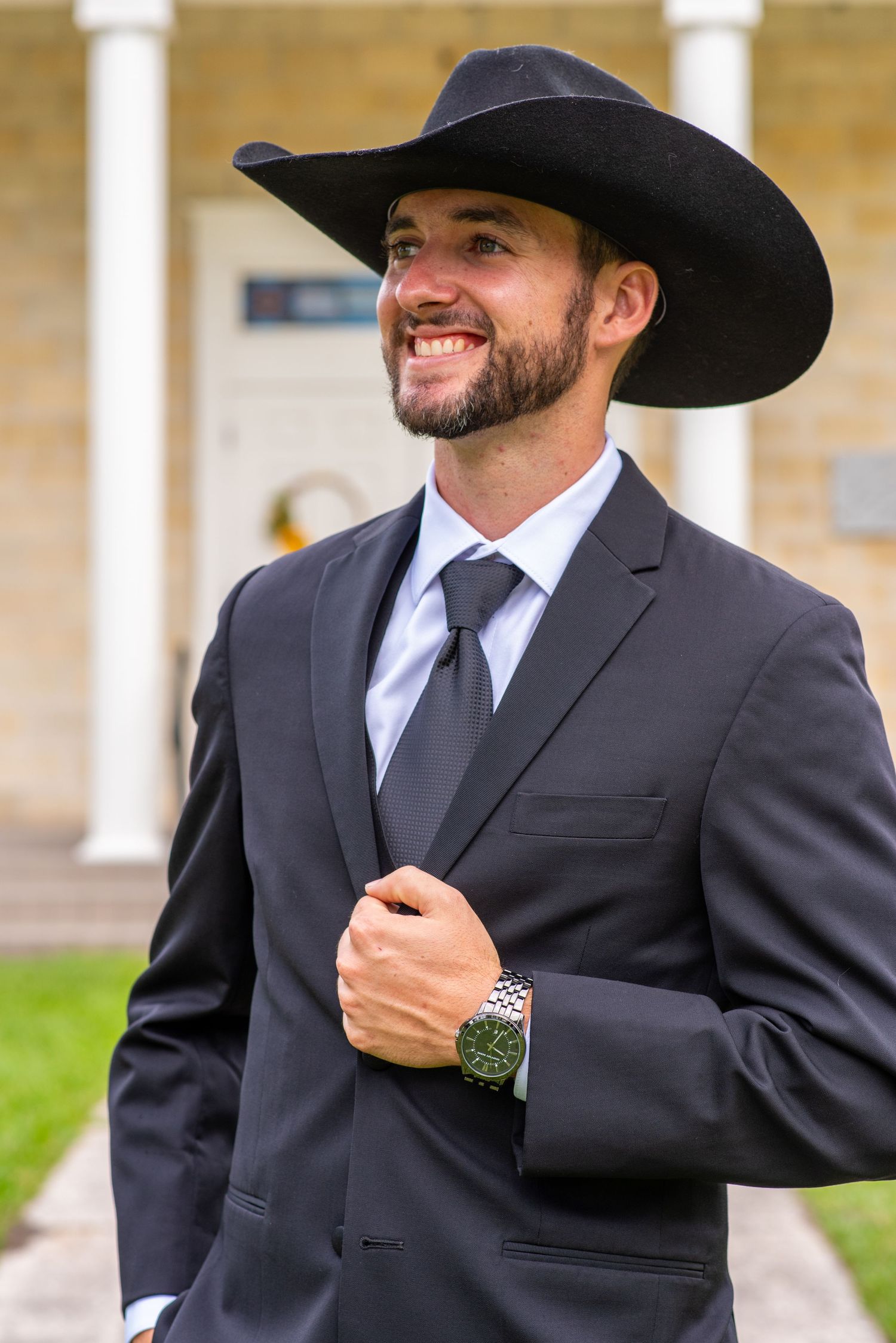 Smiling man in black suit, tie, and cowboy hat wearing a watch, standing outdoors near white columns.