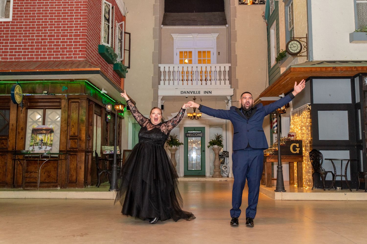 Couple in formal attire posing joyfully in a decorative indoor street set with brick buildings.